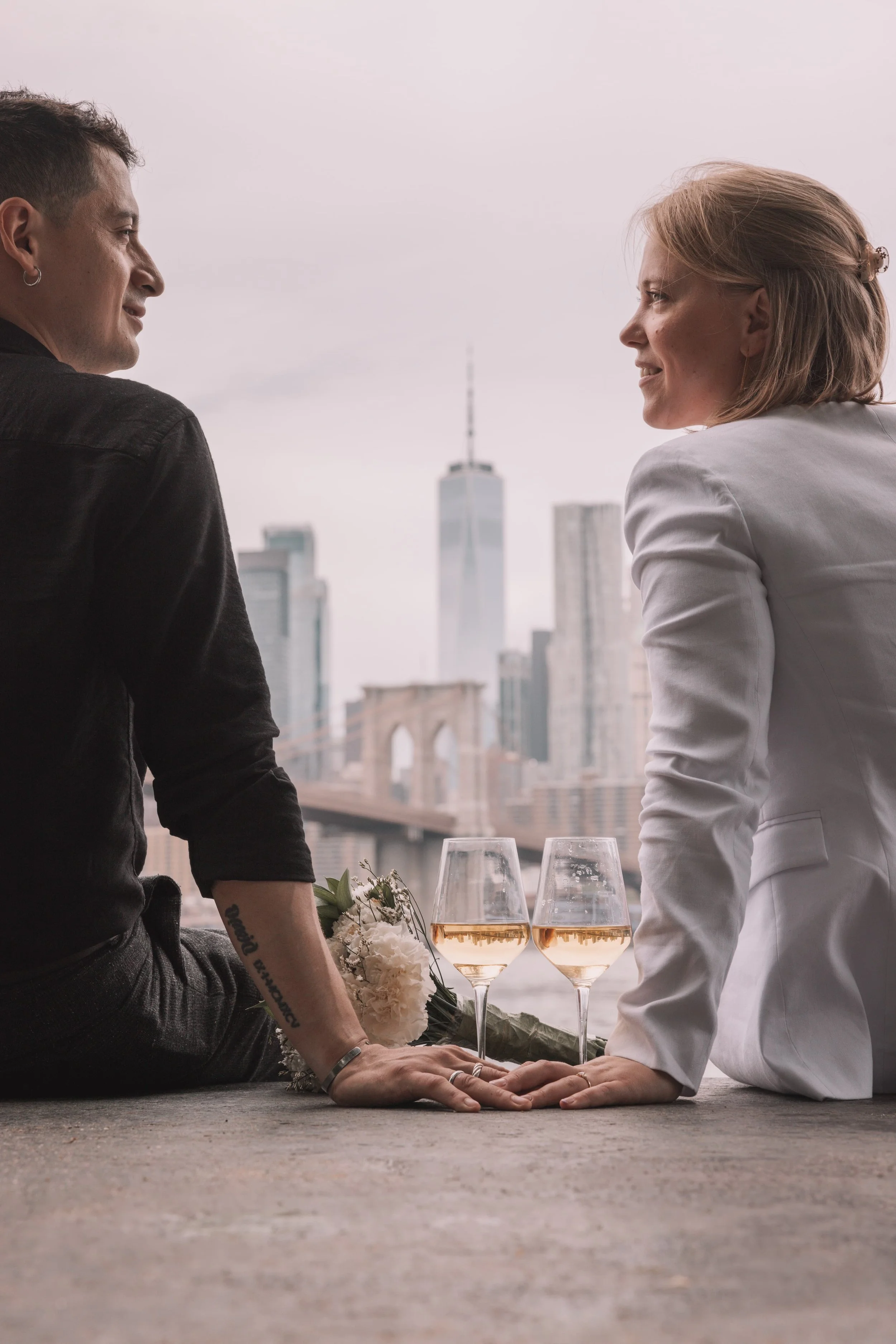 Two women sitting on the ground, holding hands, with two glasses of white wine and a bouquet of flowers, overlooking a city skyline with the Brooklyn Bridge and One World Trade Center in the background.