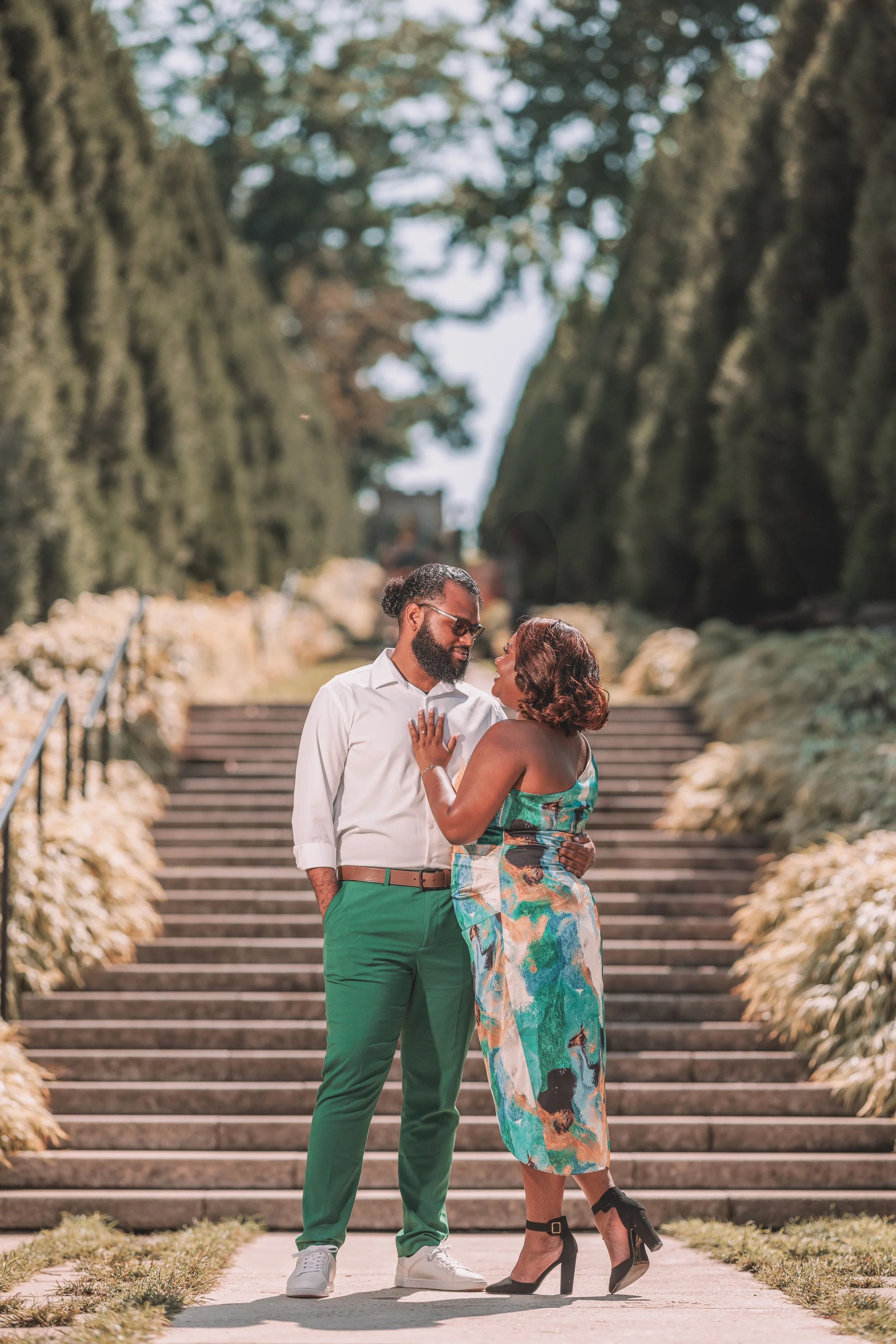 A couple standing on steps outdoors, embracing and looking at each other, surrounded by greenery and trees.