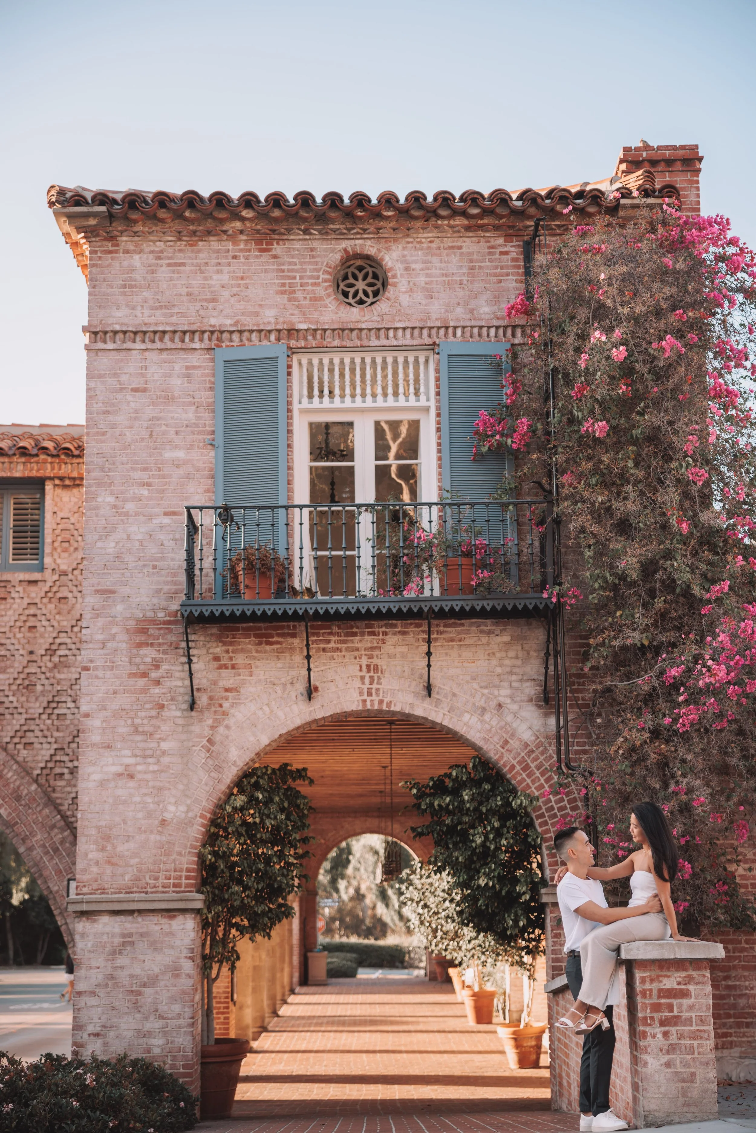 A couple in white clothing sitting on a brick ledge in front of a pink brick building with an arched passageway, potted plants, and a flowering vine, during the daytime.