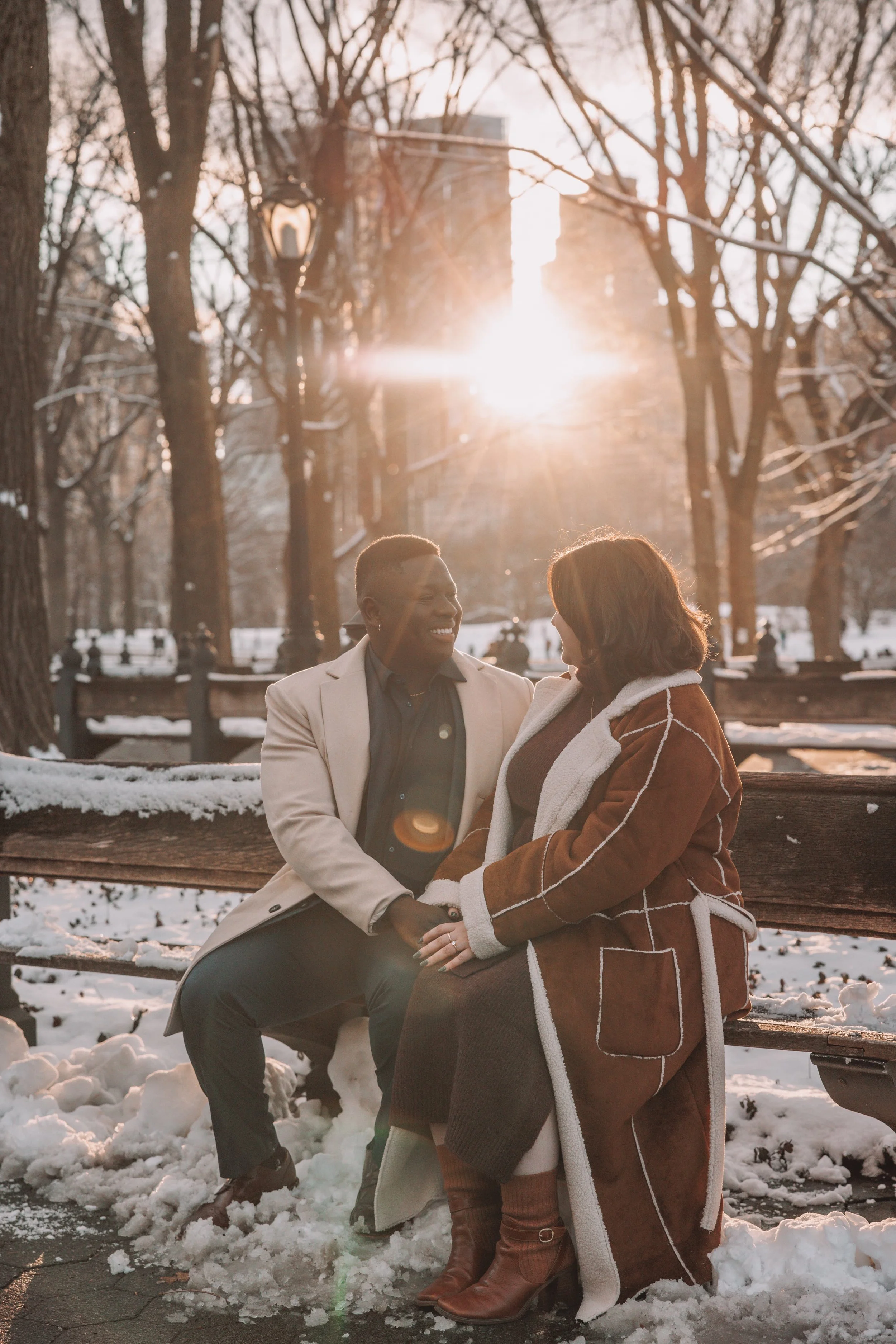 A couple sitting on a park bench in winter, holding hands and smiling at each other, with snow on the ground and blooming trees in the background, in front of the setting sun.