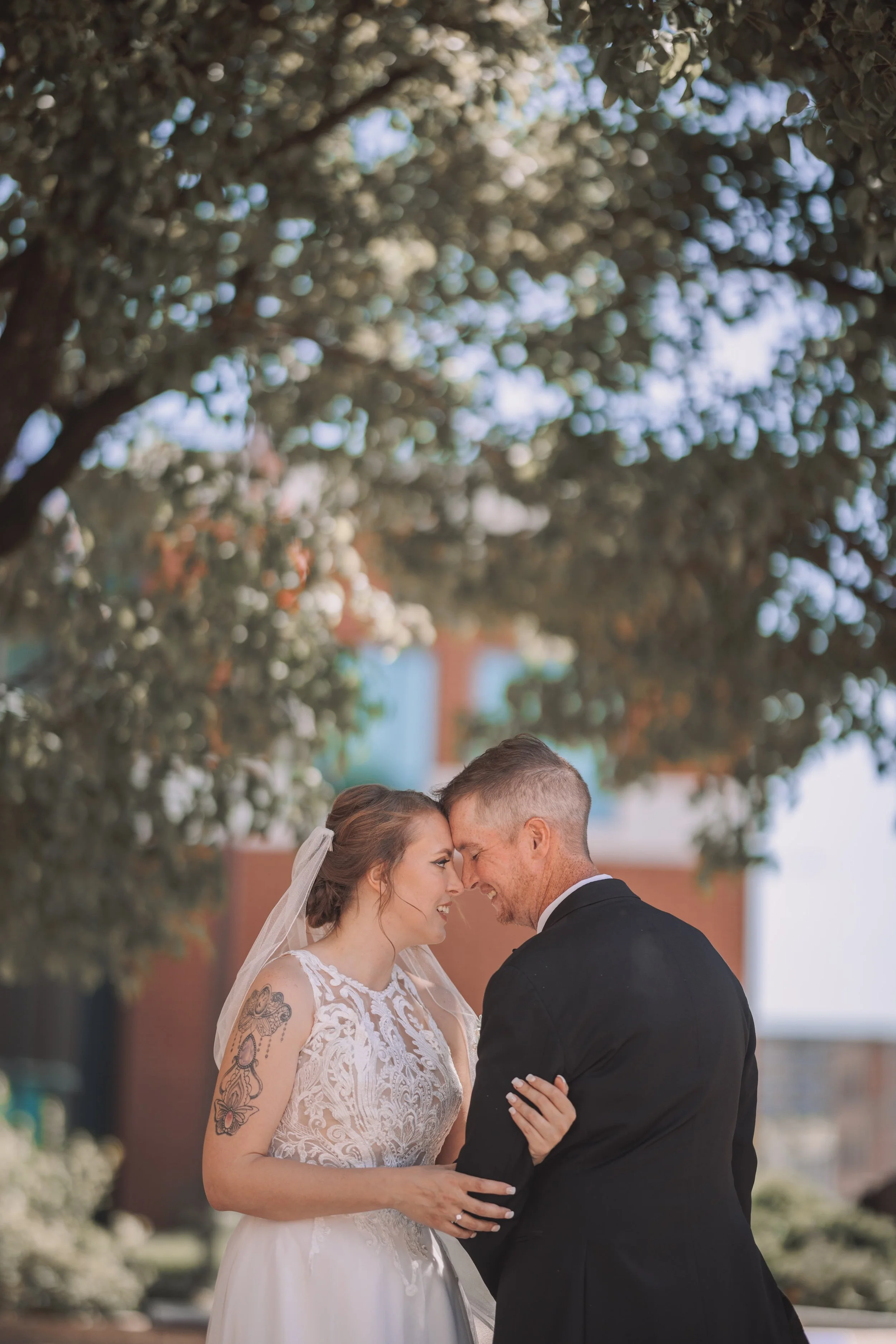 A bride and groom sharing a close, joyful moment outdoors during their wedding, with their foreheads touching and smiling.