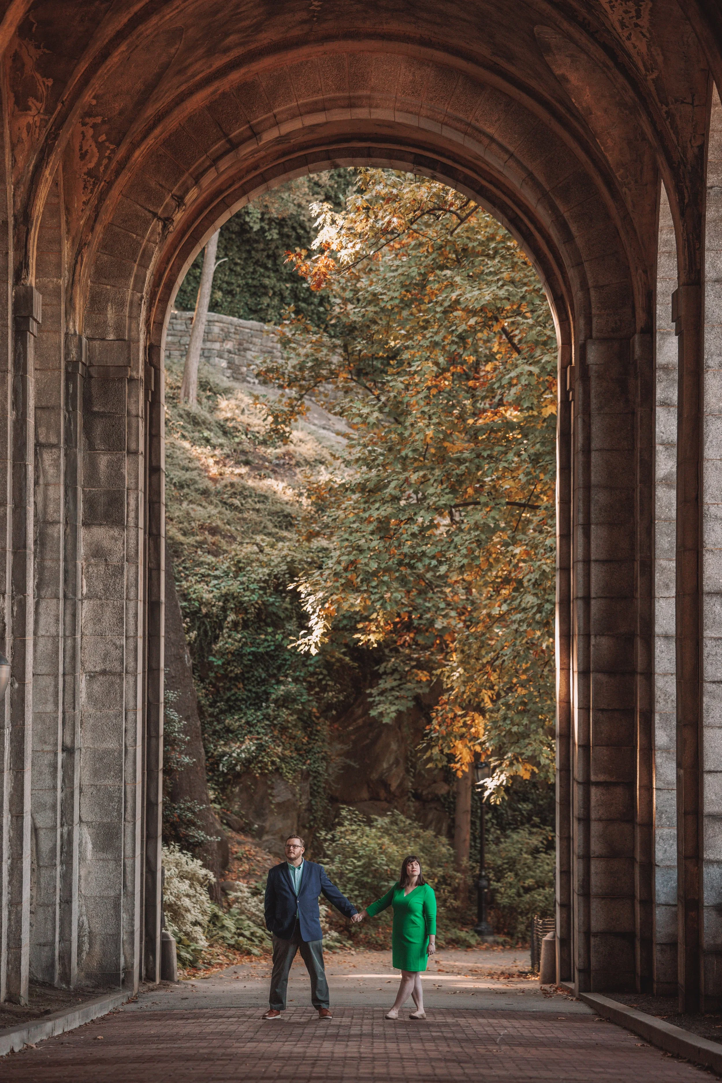 A man and woman holding hands under a large stone archway with autumn trees in the background.