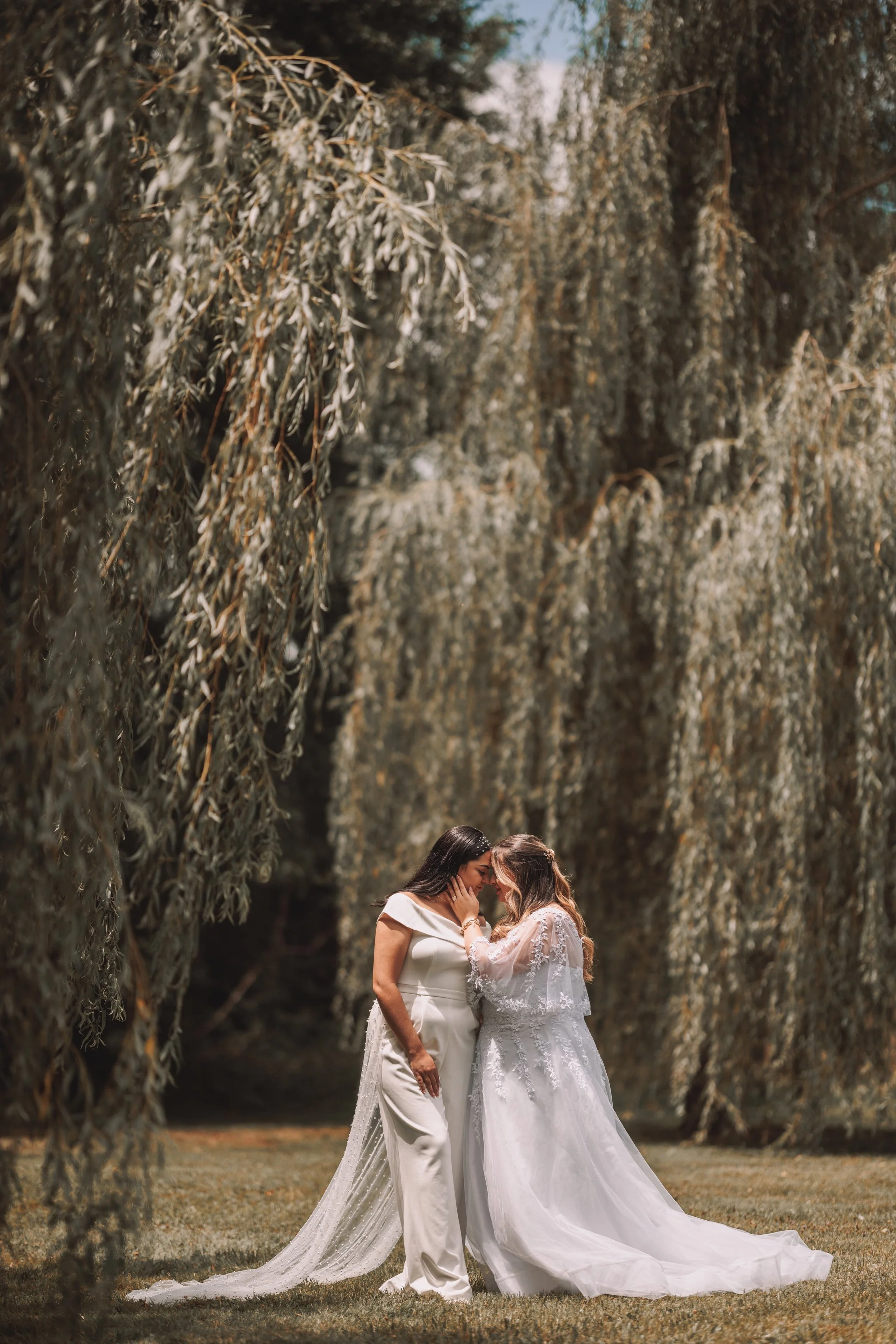 Two women in wedding dresses sharing a tender moment under hanging willow trees outdoors.