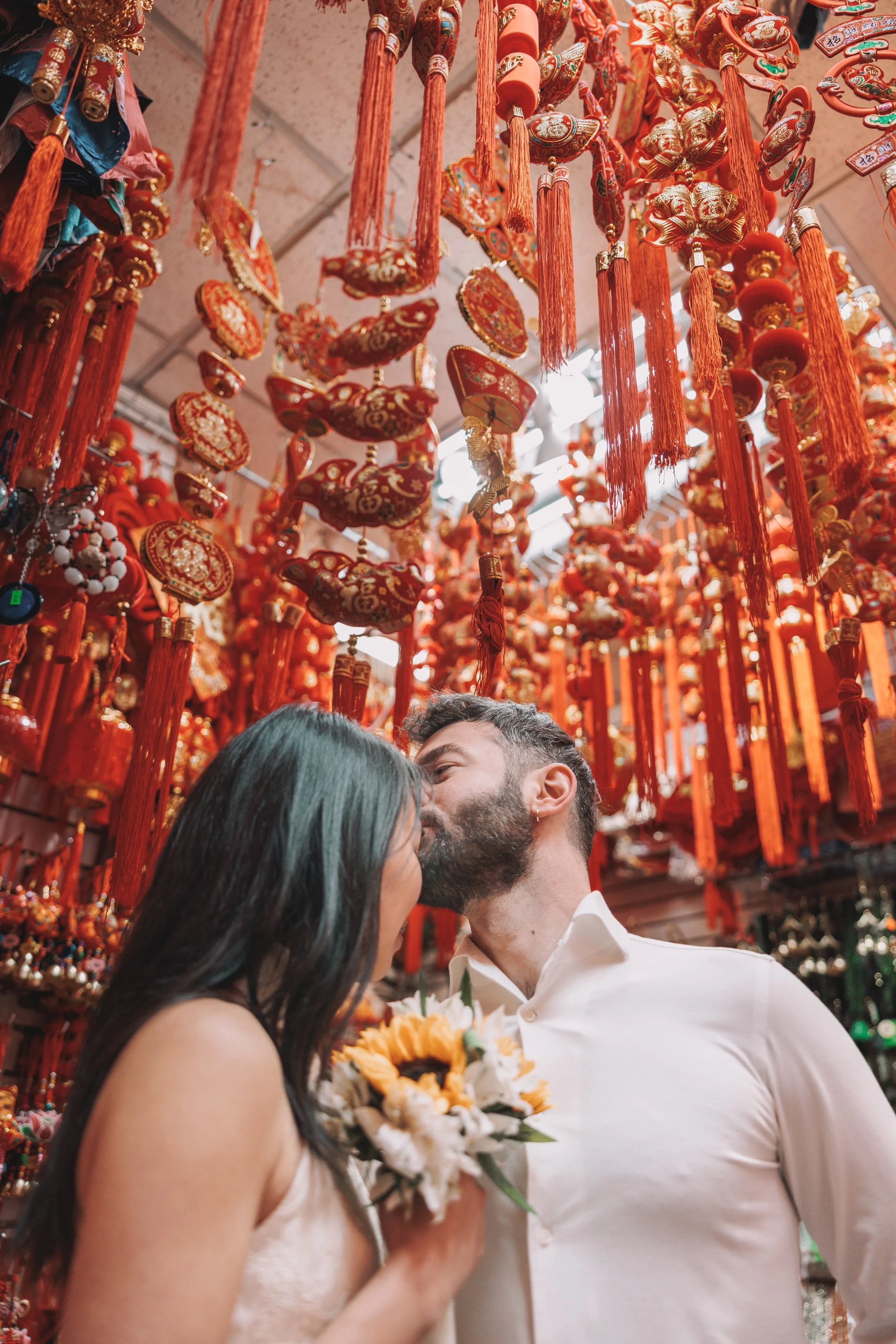 A newlywed couple nose to nose in a shop filled with red and gold Chinese New Year decorations.