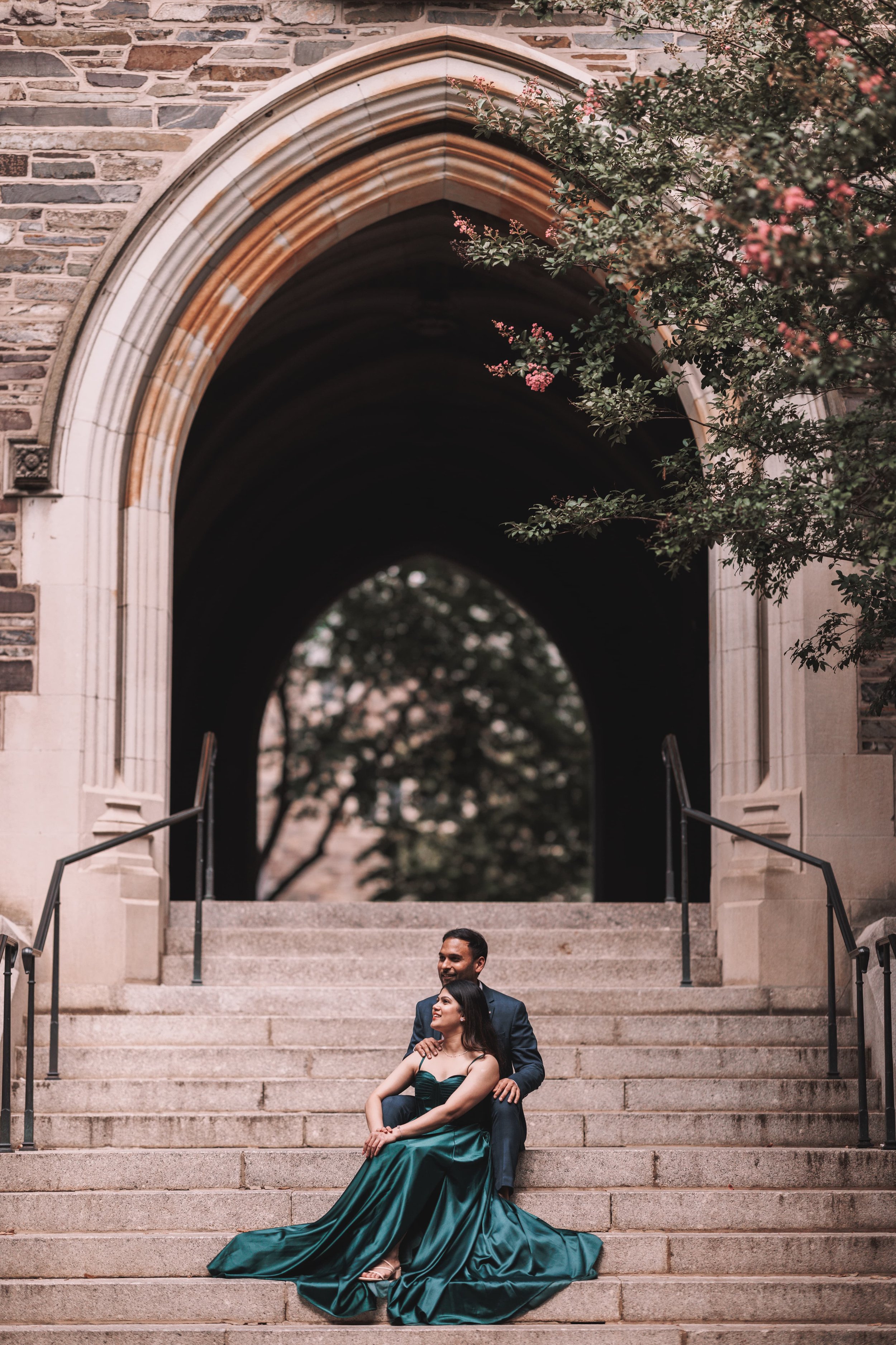 A couple sitting on steps in front of a church arch. The woman is wearing a long, dark green satin gown and the man is dressed in a dark suit. They are smiling and looking at each other, surrounded by trees and stone temple details.