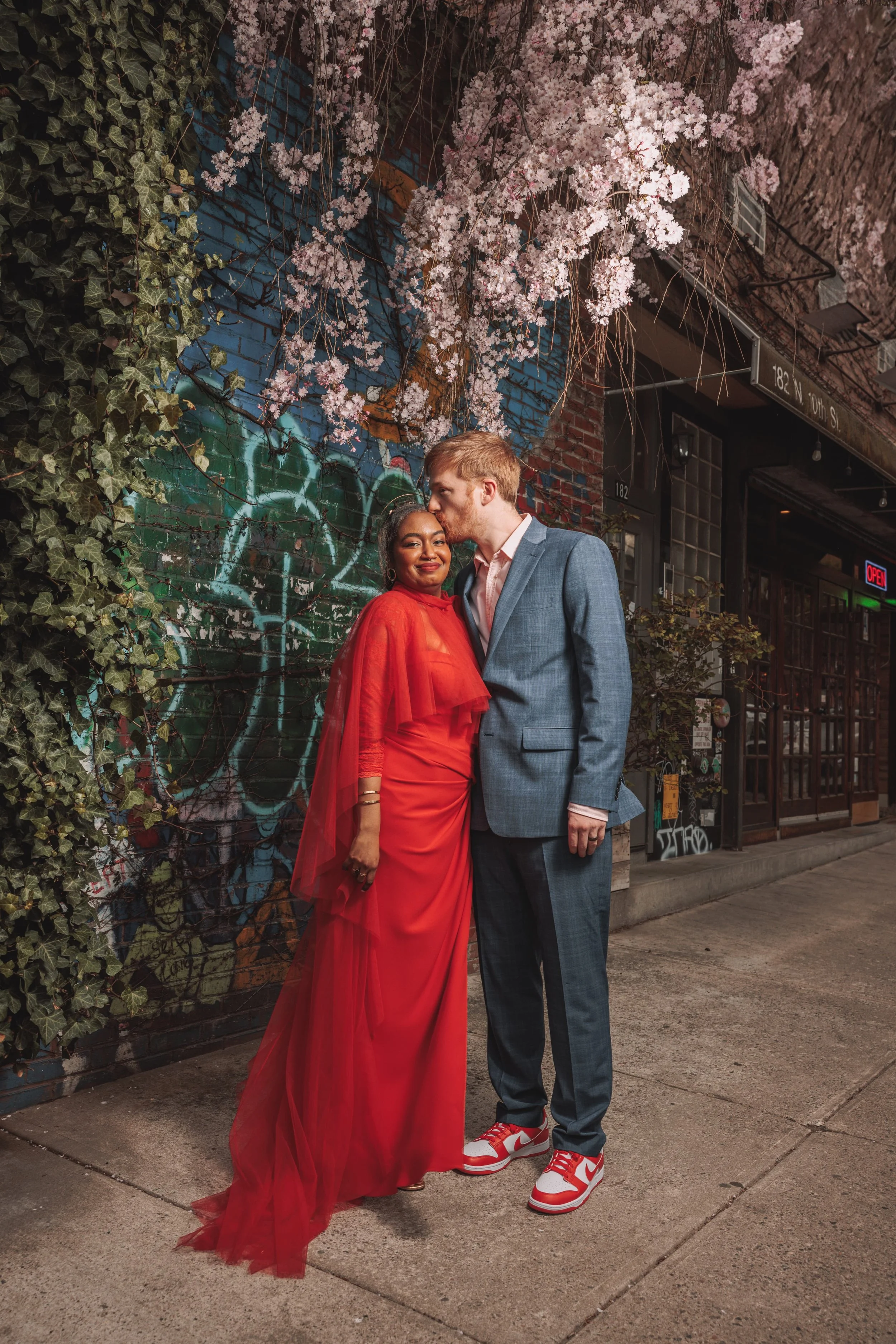 A couple standing on a city sidewalk at night, with a graffiti-covered wall and flowering tree behind them. The woman is wearing a red dress with sheer flowing fabric, smiling at the camera, and the man is dressed in a gray suit with a pink shirt, ki
