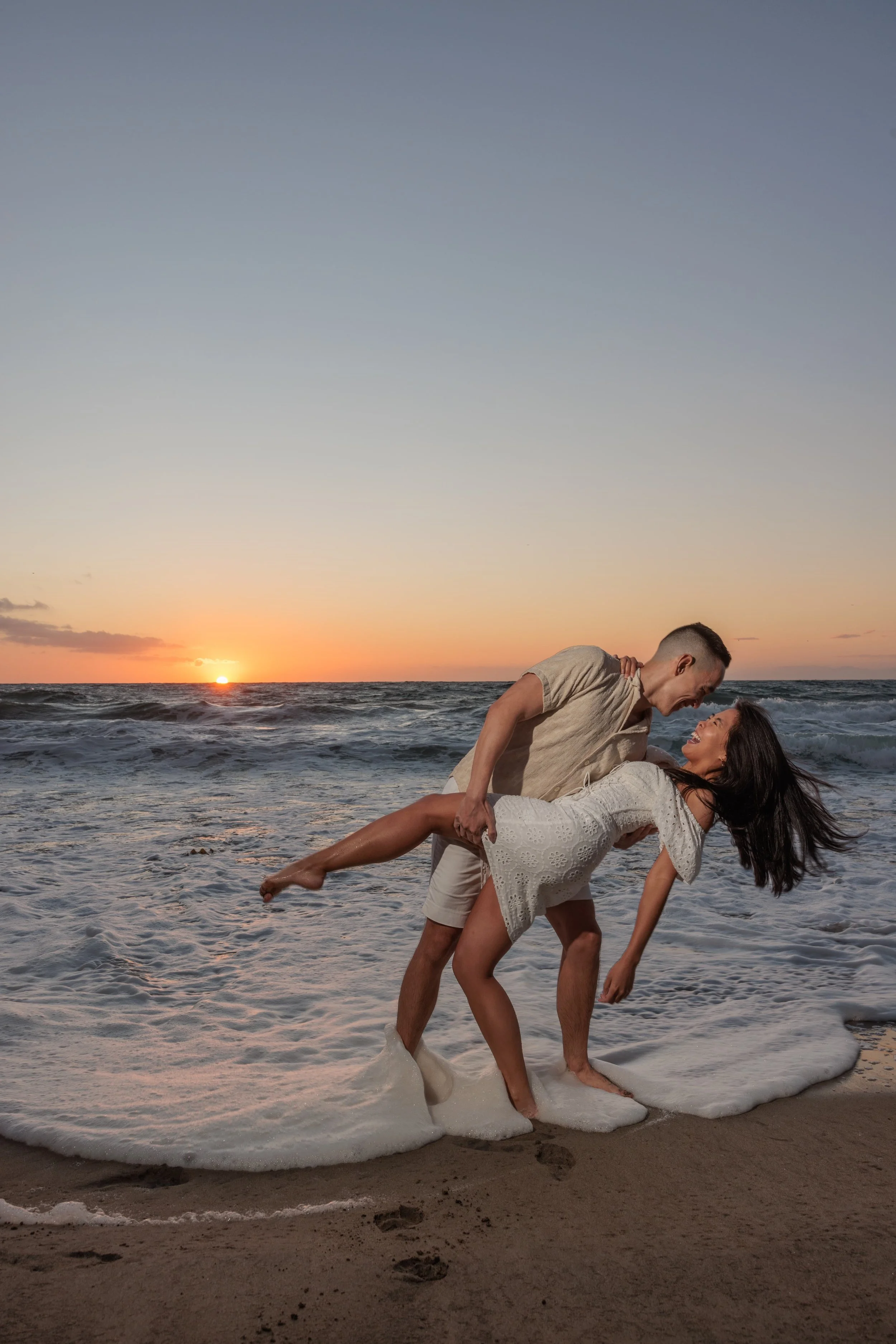 A couple dancing in the water at sunset on the beach, with waves and a colorful sky in the background.