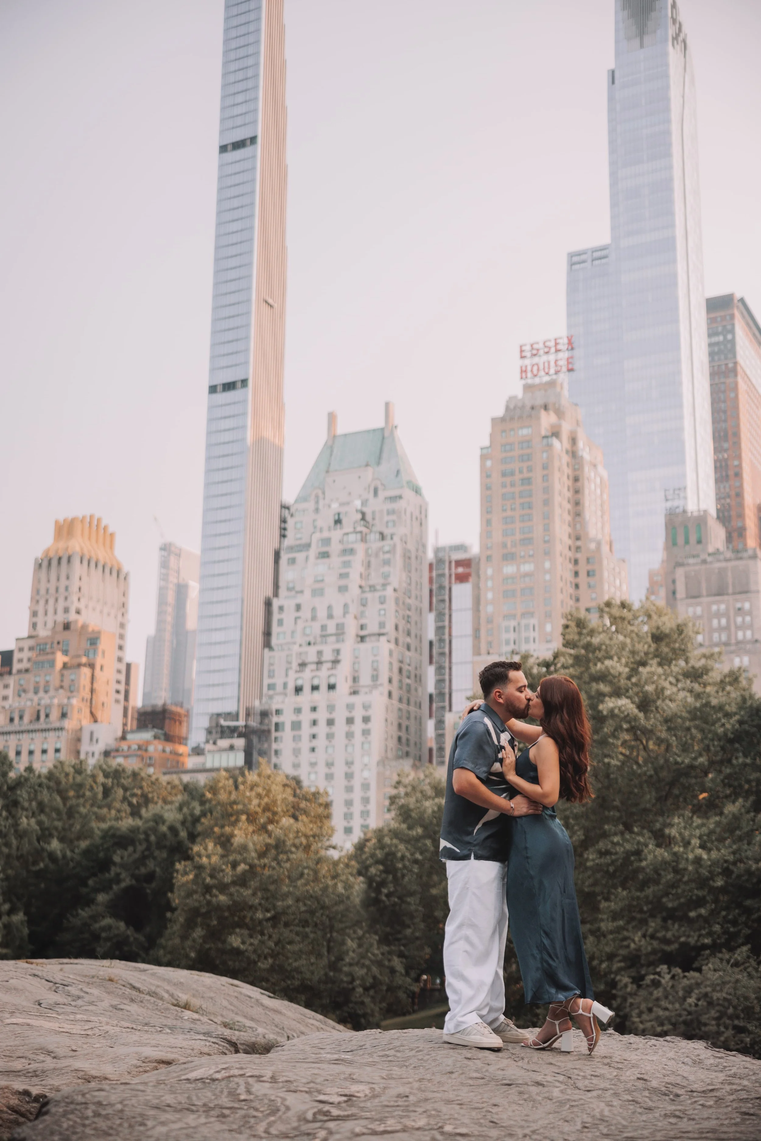 A couple sharing a kiss while standing on a large rock in a park with New York City skyscrapers in the background.