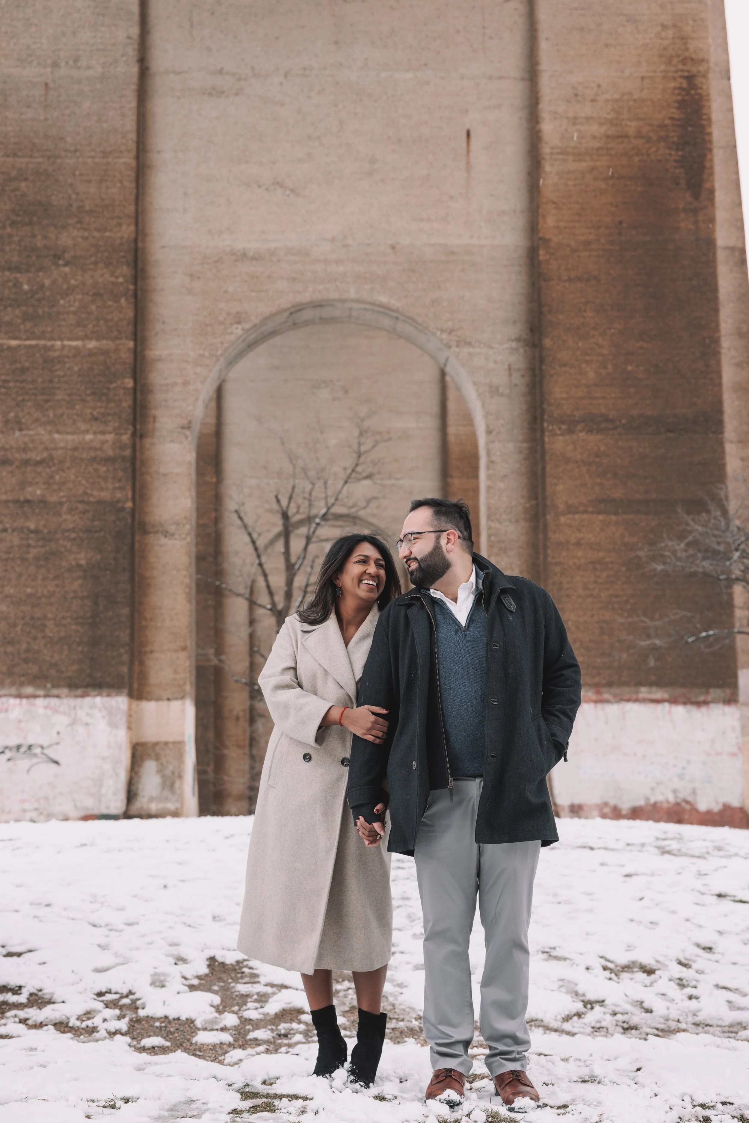 A smiling couple walking hand in hand on a snowy ground in front of a large brick wall with a tree in the background.