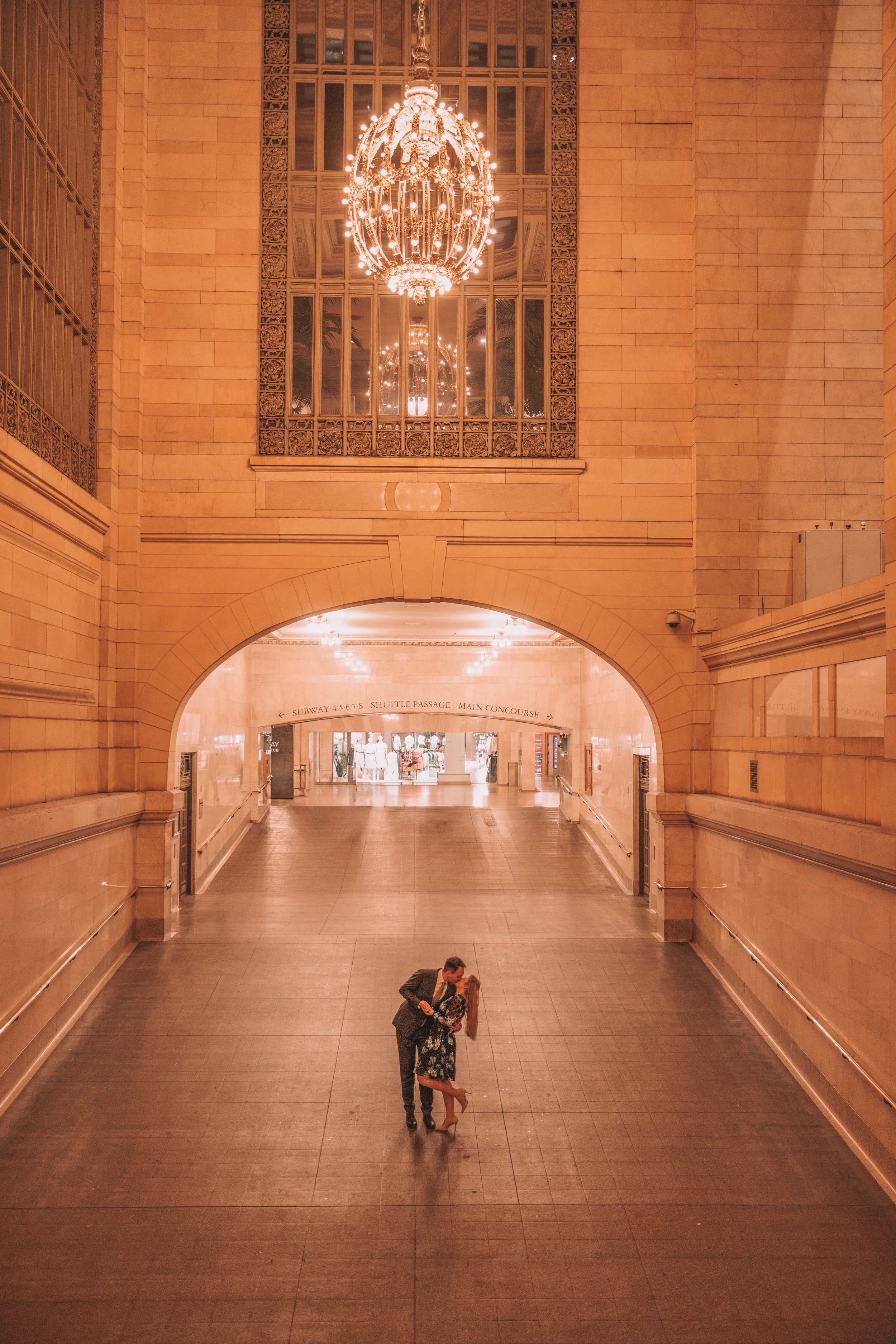 A couple dancing in a grand, well-lit indoor space with high ceilings, chandeliers, and large windows.