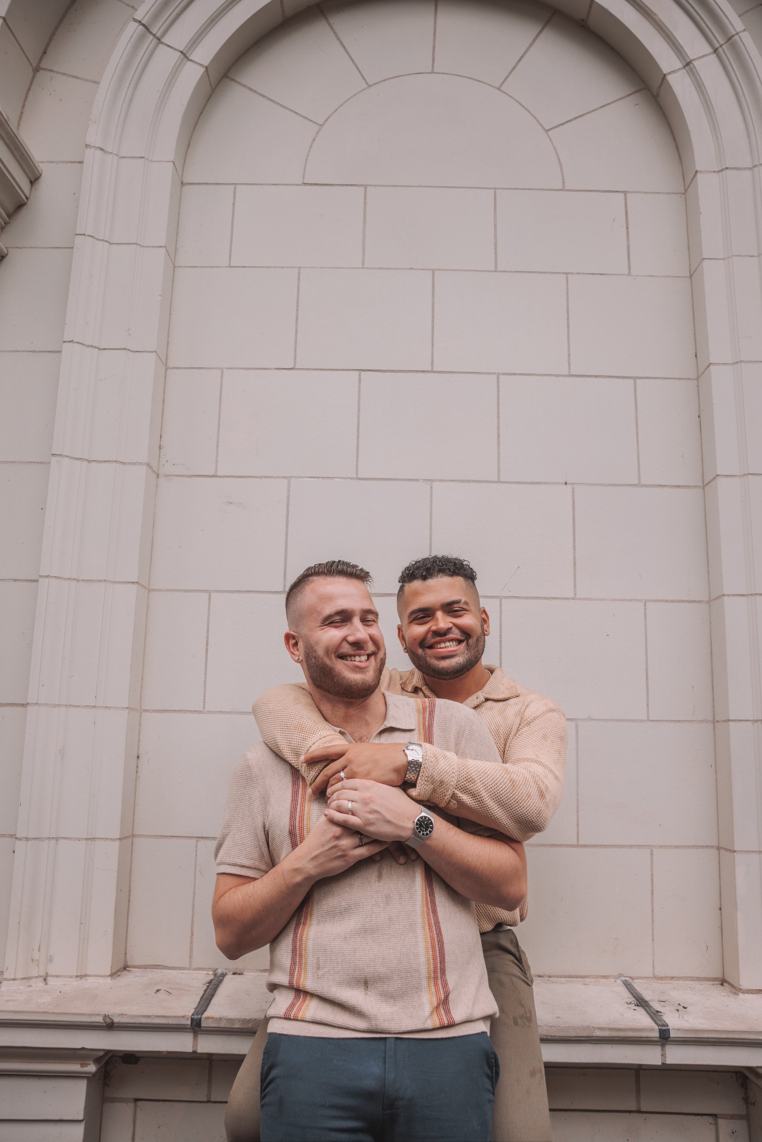 Two men smiling and hugging in front of a beige stone wall with an arched design.
