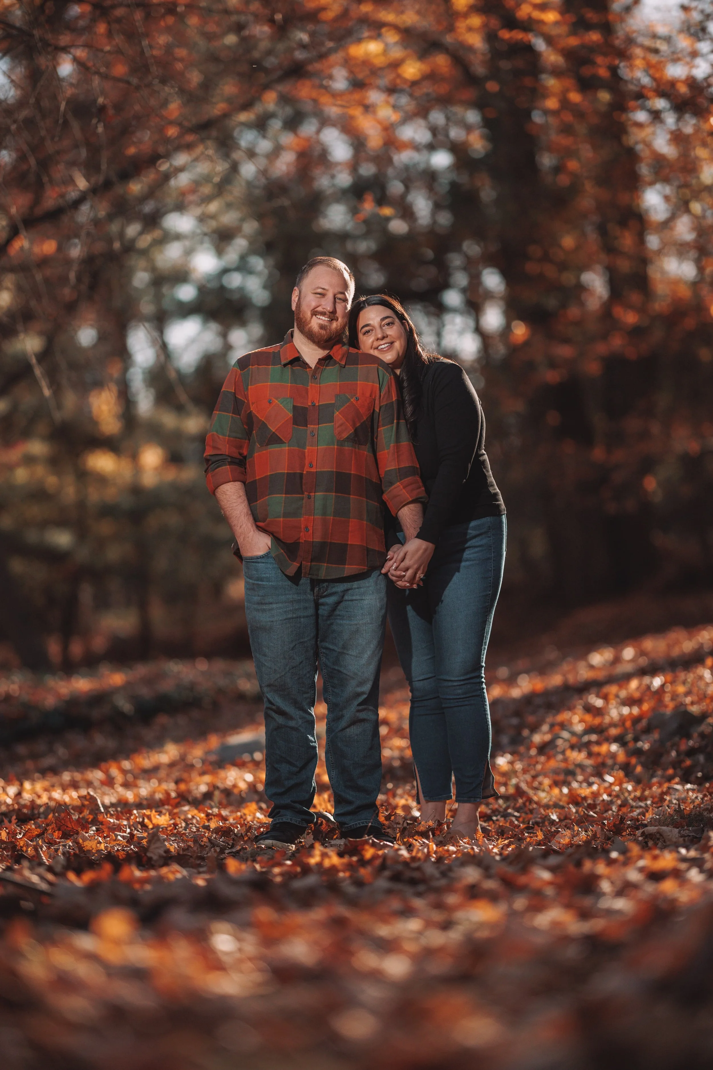 A smiling couple standing hand in hand on a fallen leaf-covered path during fall, surrounded by trees with orange and brown leaves.