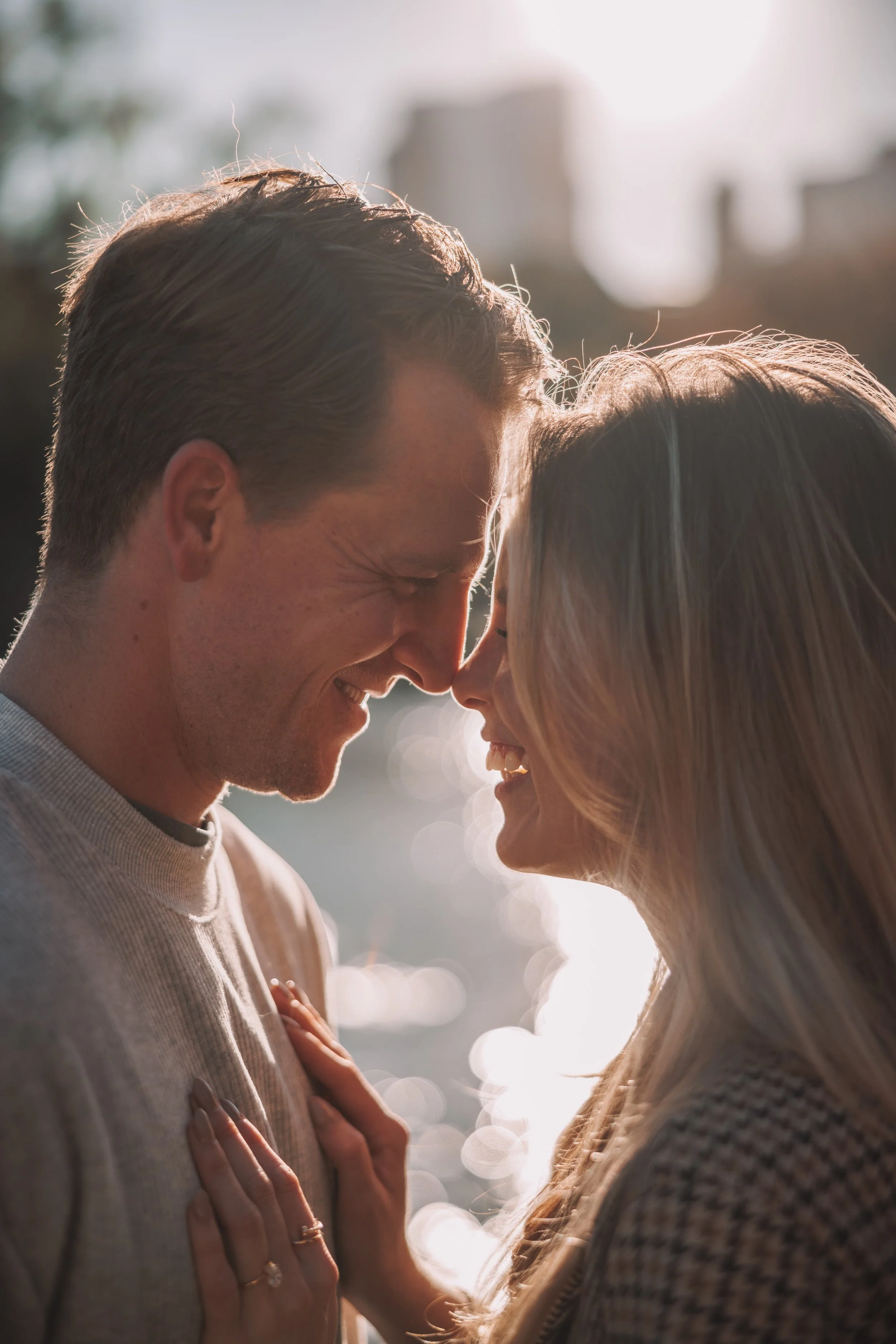 A close-up of a couple smiling with foreheads and noses touching, backlit by sunlight, outdoors with water and city buildings in the background.