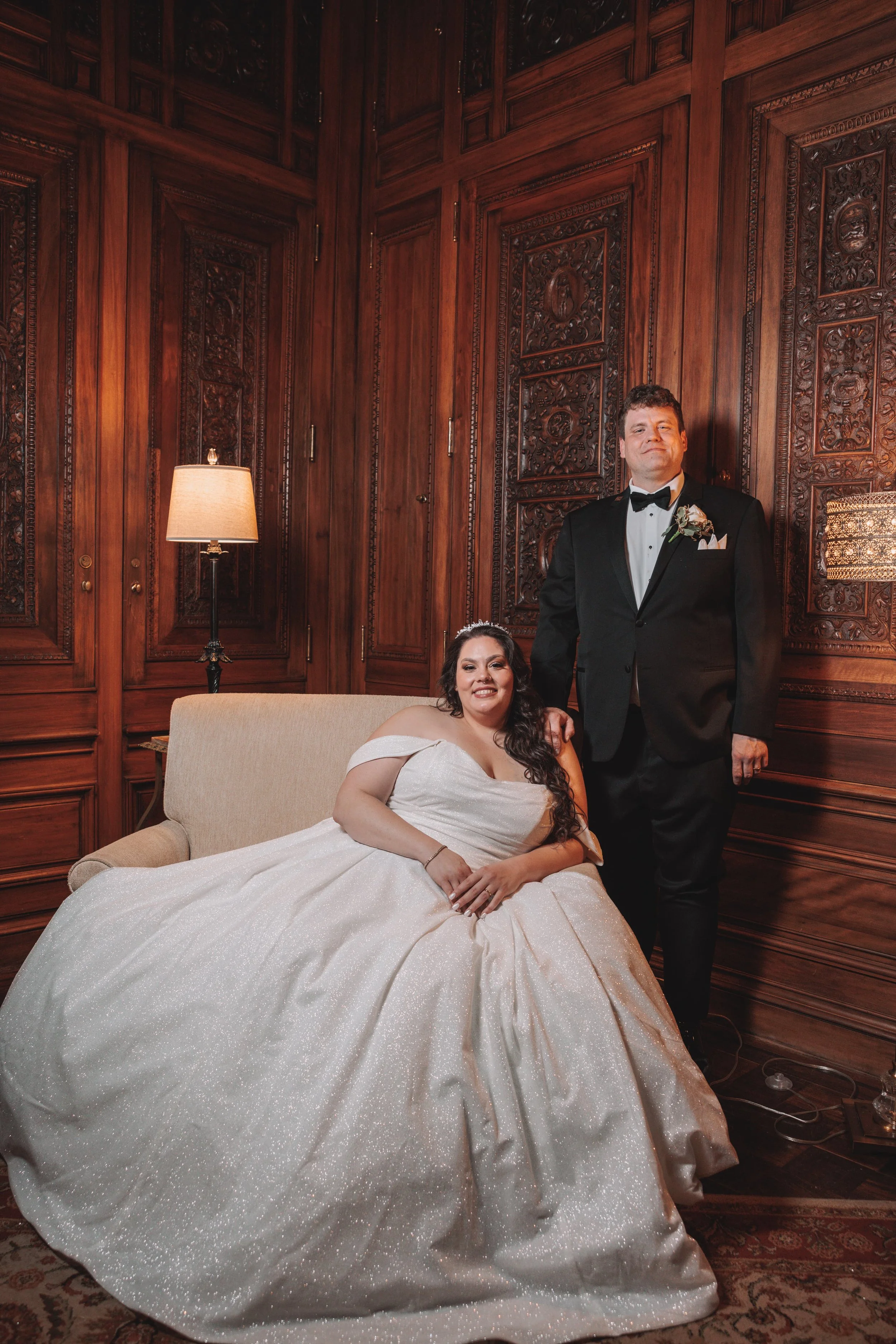 A bride and groom in formal wedding attire, posing inside a wood-paneled room. The bride is seated on a beige couch, wearing a white gown, and the groom is standing next to her in a black tuxedo.