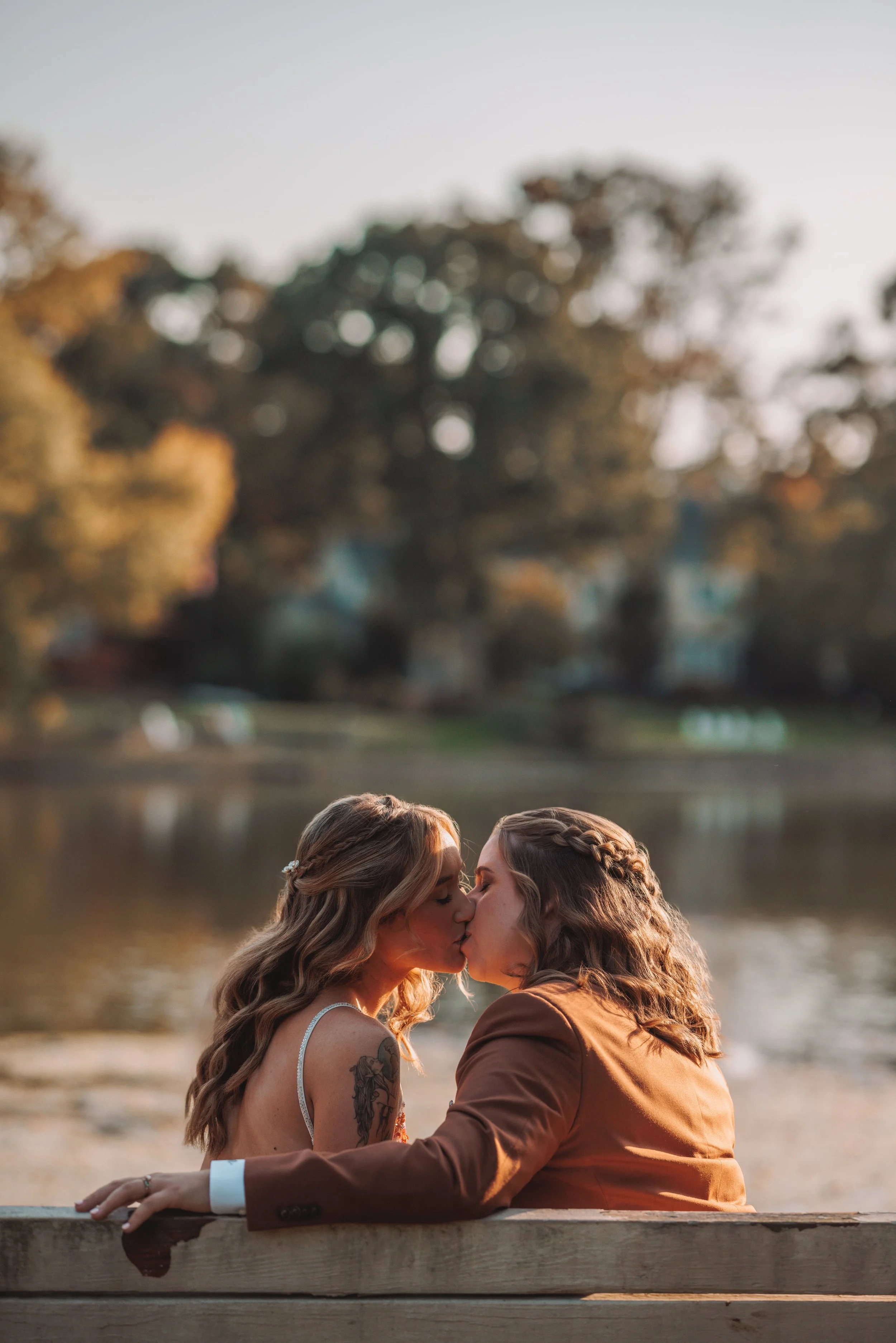Two women kissing on a park bench by a river, with trees and houses in the background during sunset.