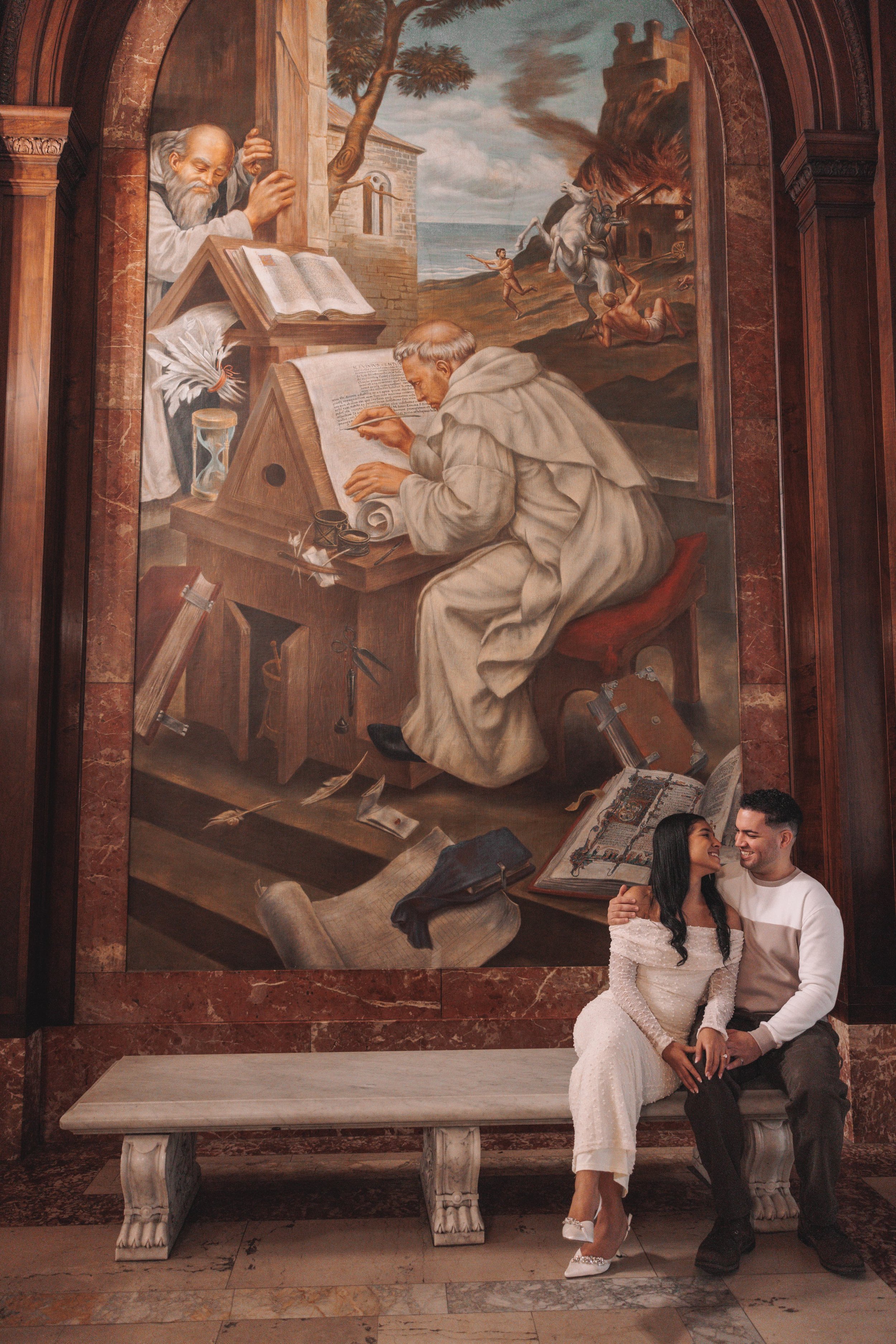 A couple sits on a marble bench in front of a large religious painting that depicts a historical or biblical scene of a scholar writing at a desk, with a saintly figure reading a book nearby, and a biblical scene with a horseman and a woman in the ba