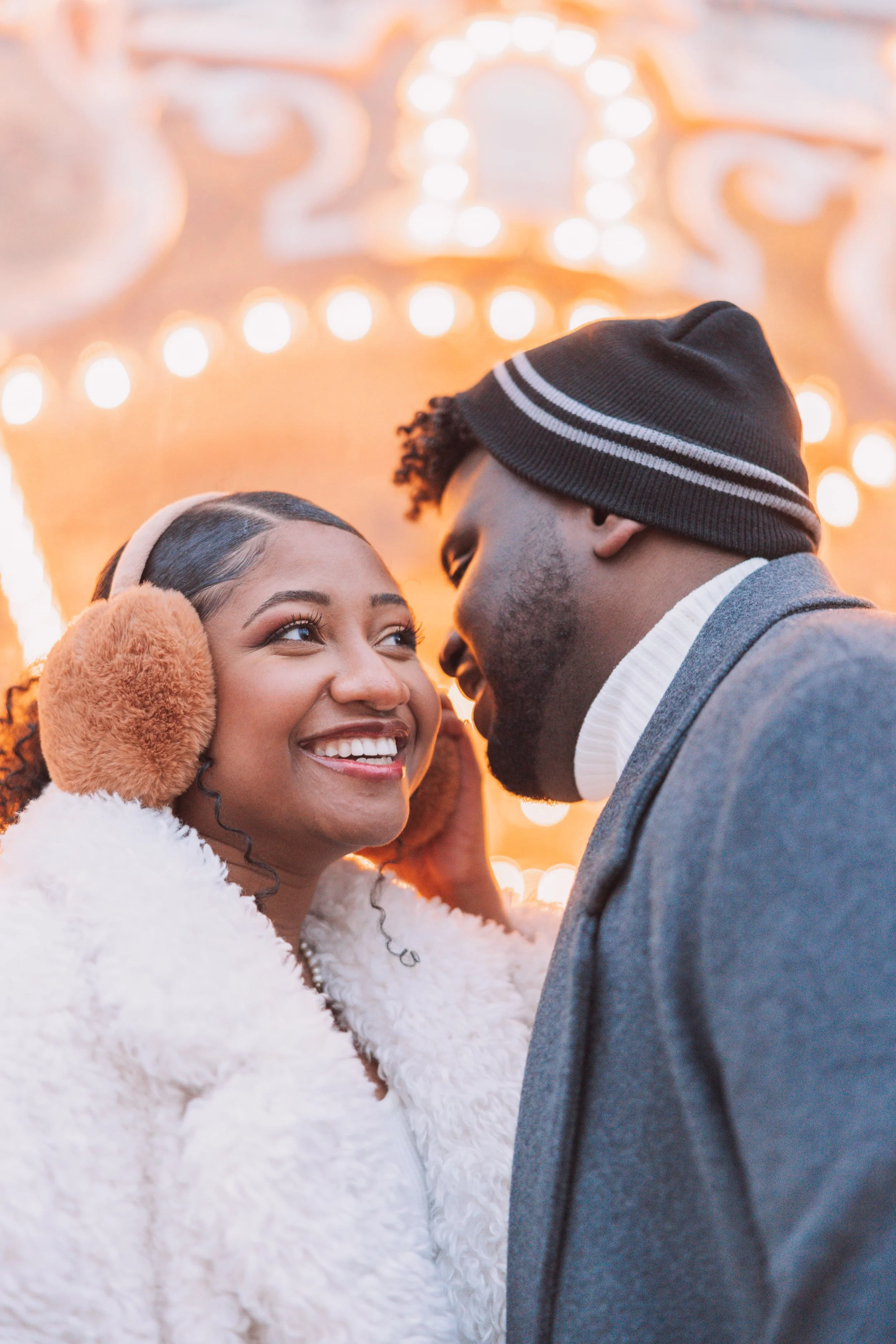 A smiling woman with curly hair and earmuffs, and a man wearing a beanie hat, sharing an intimate moment at a festive, lit-up outdoor event.