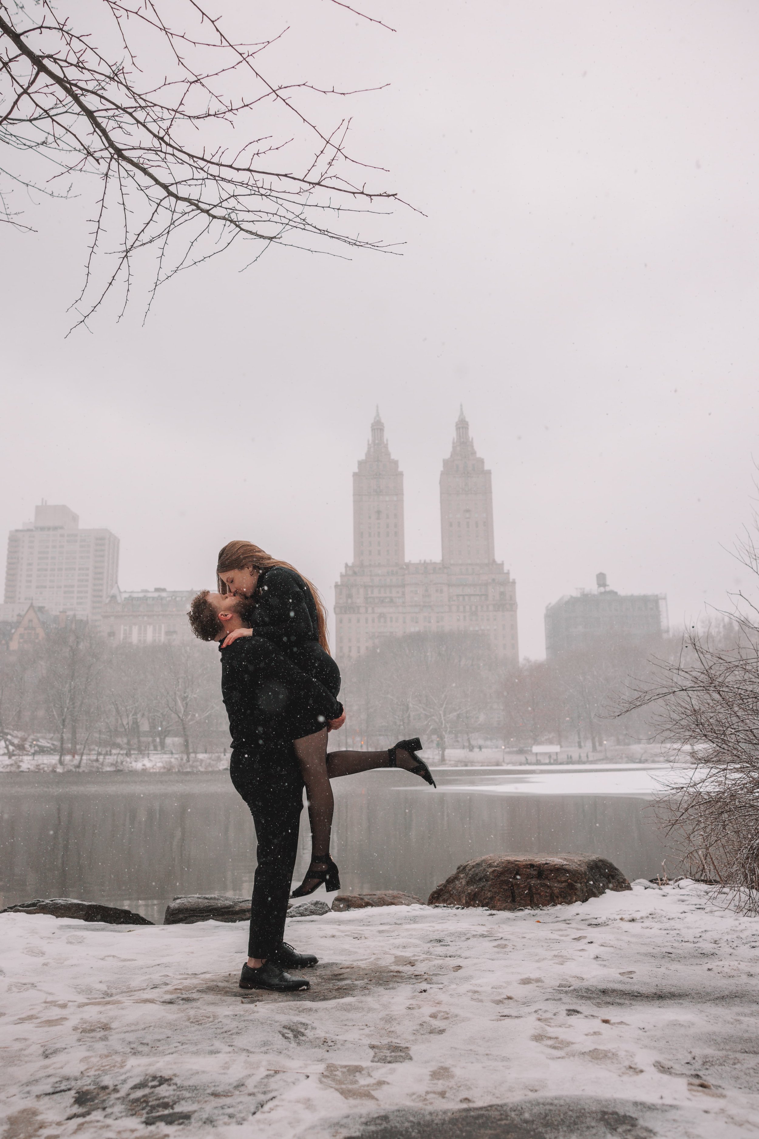 A couple in love in a snowy park, with the city skyline in the background, the man lifting the woman as they kiss.