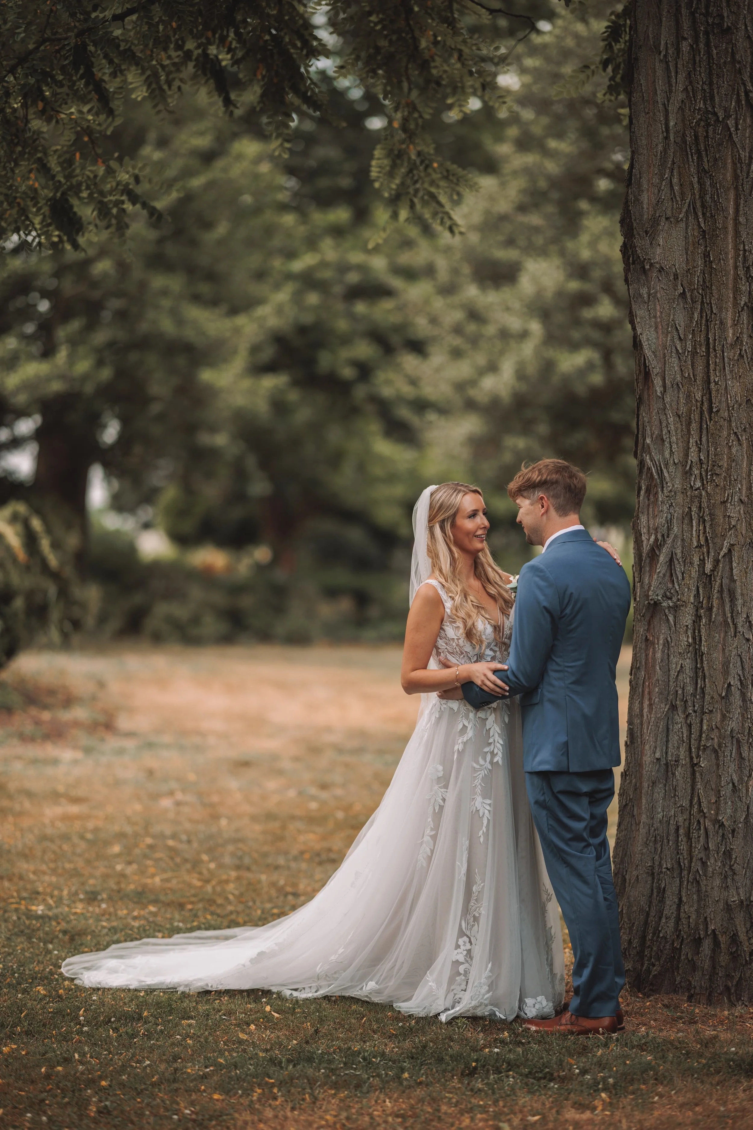 A bride and groom stand close together near a big tree in a park, sharing a kiss or intimate moment, surrounded by greenery.