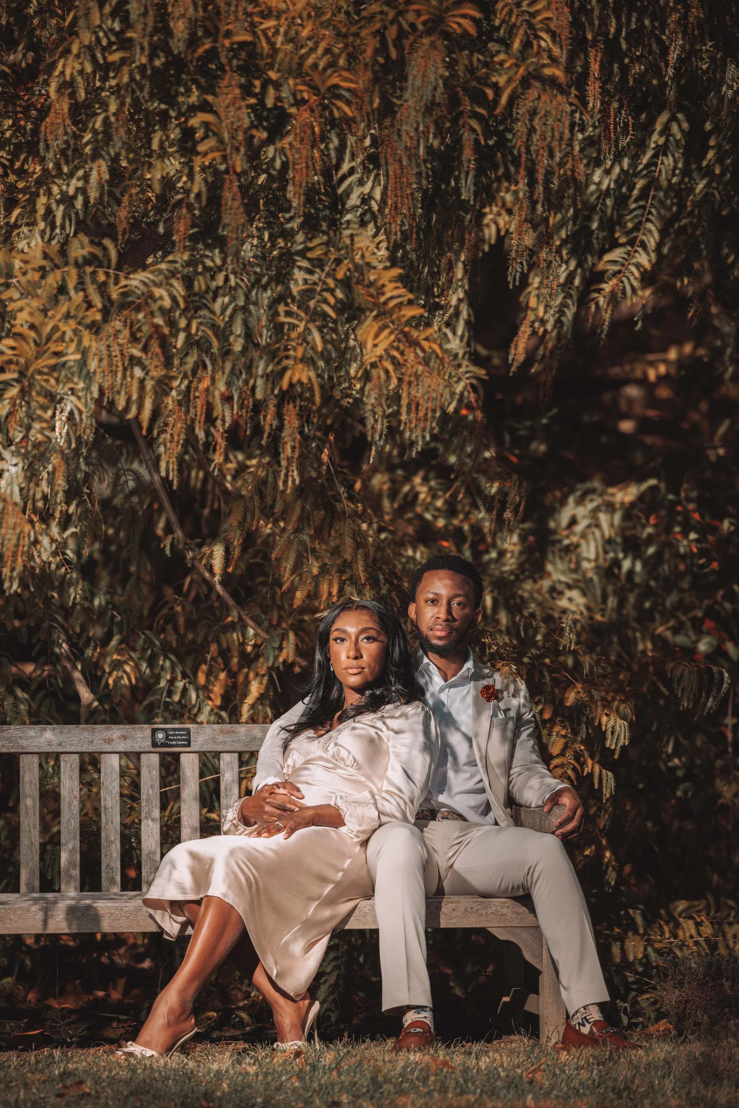 A couple sitting on a wooden park bench at night with large trees in the background. The woman is in a shiny beige dress, and the man is in a light-colored suit with a floral lapel pin.