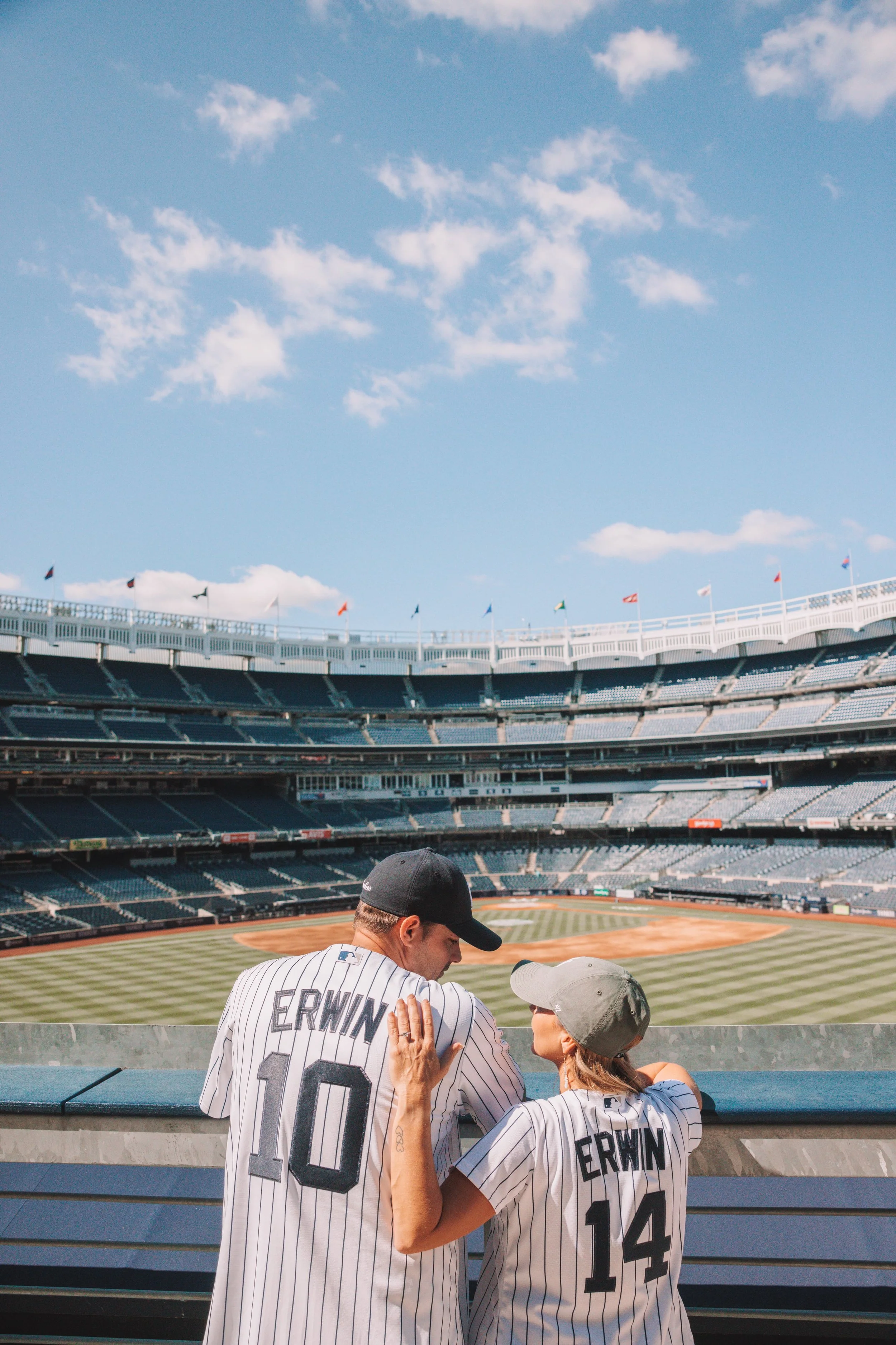 A man and woman wearing New York Yankees baseball jerseys standing at a stadium, talking and leaning on a railing with an empty baseball field and blue sky in the background.
