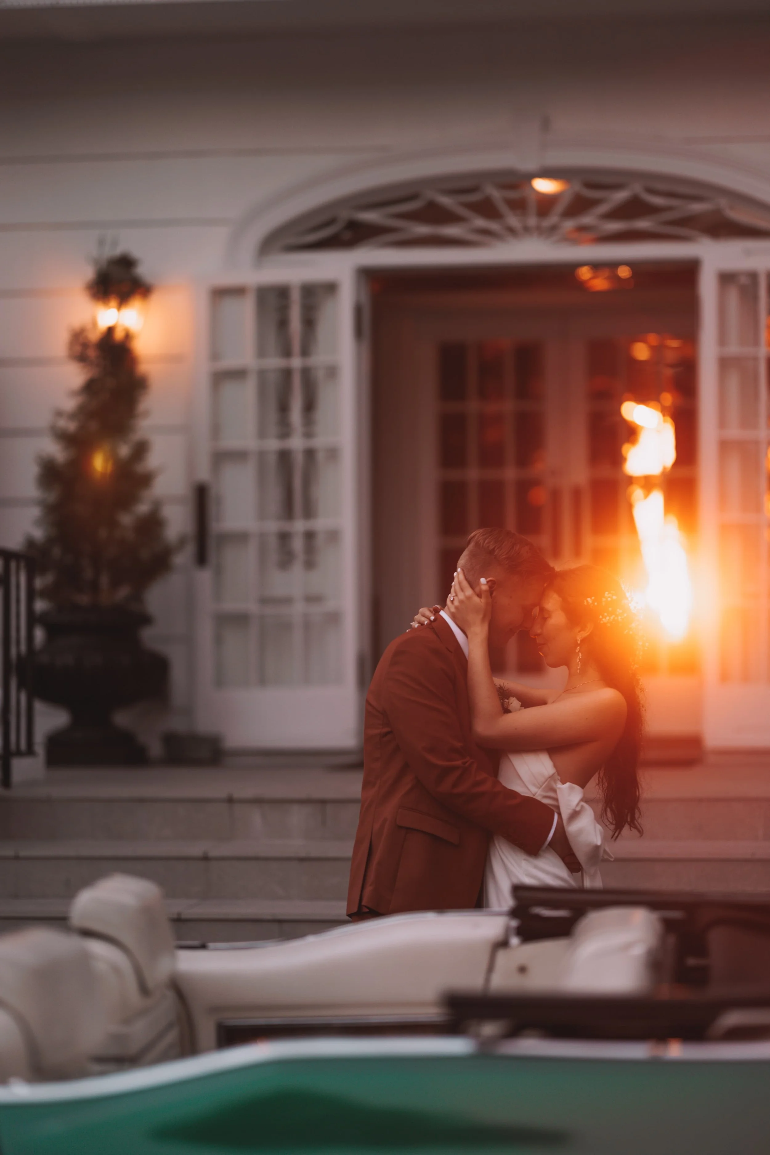 A couple dressed in wedding attire embracing in front of a house with open doors. The sunset light creates a warm glow and a flare in the background. A boat is visible in the foreground.