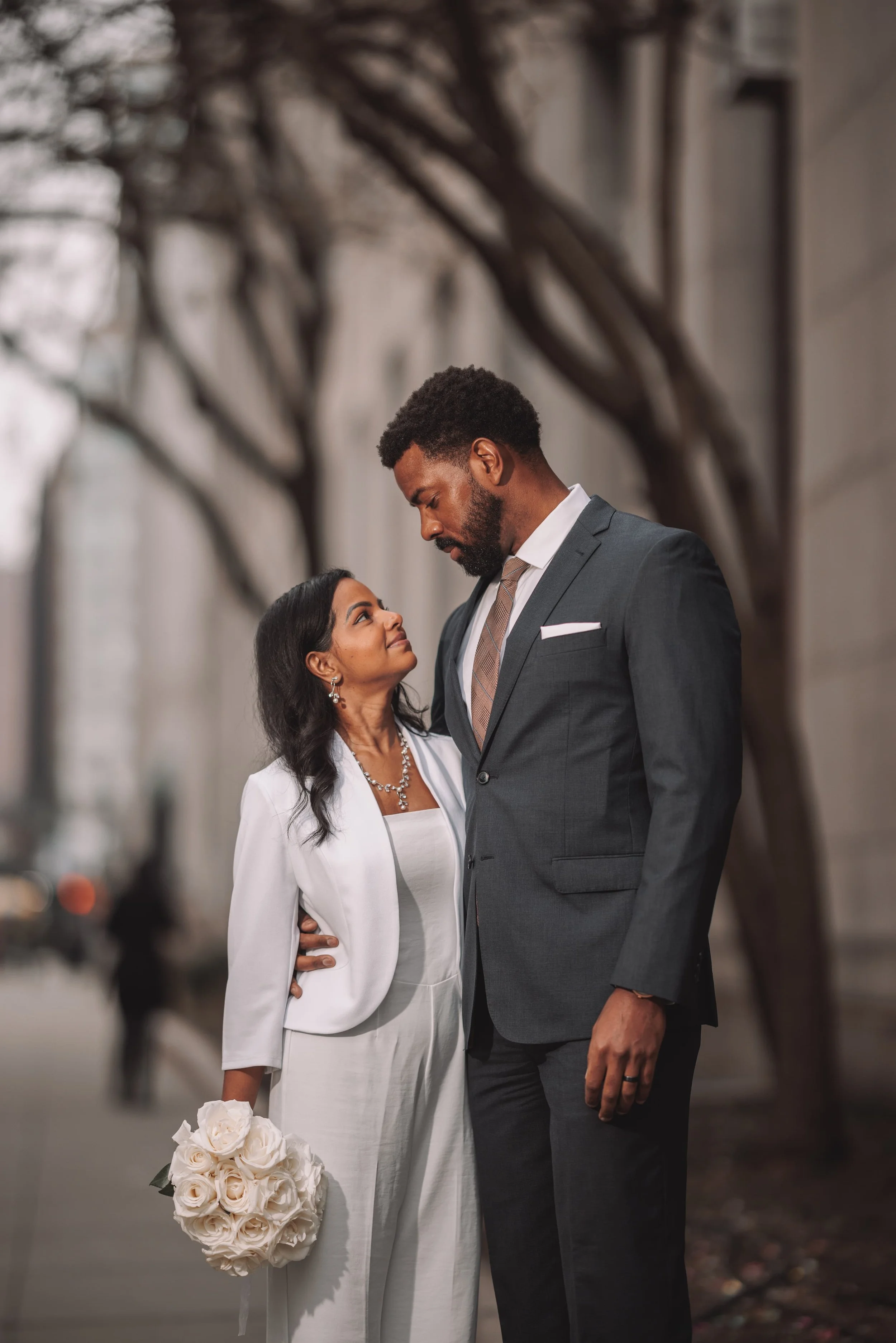 A woman in white holding a bouquet of white roses looking up at a tall man in a gray suit looking down at her in an outdoor urban setting with leafless trees in the background.