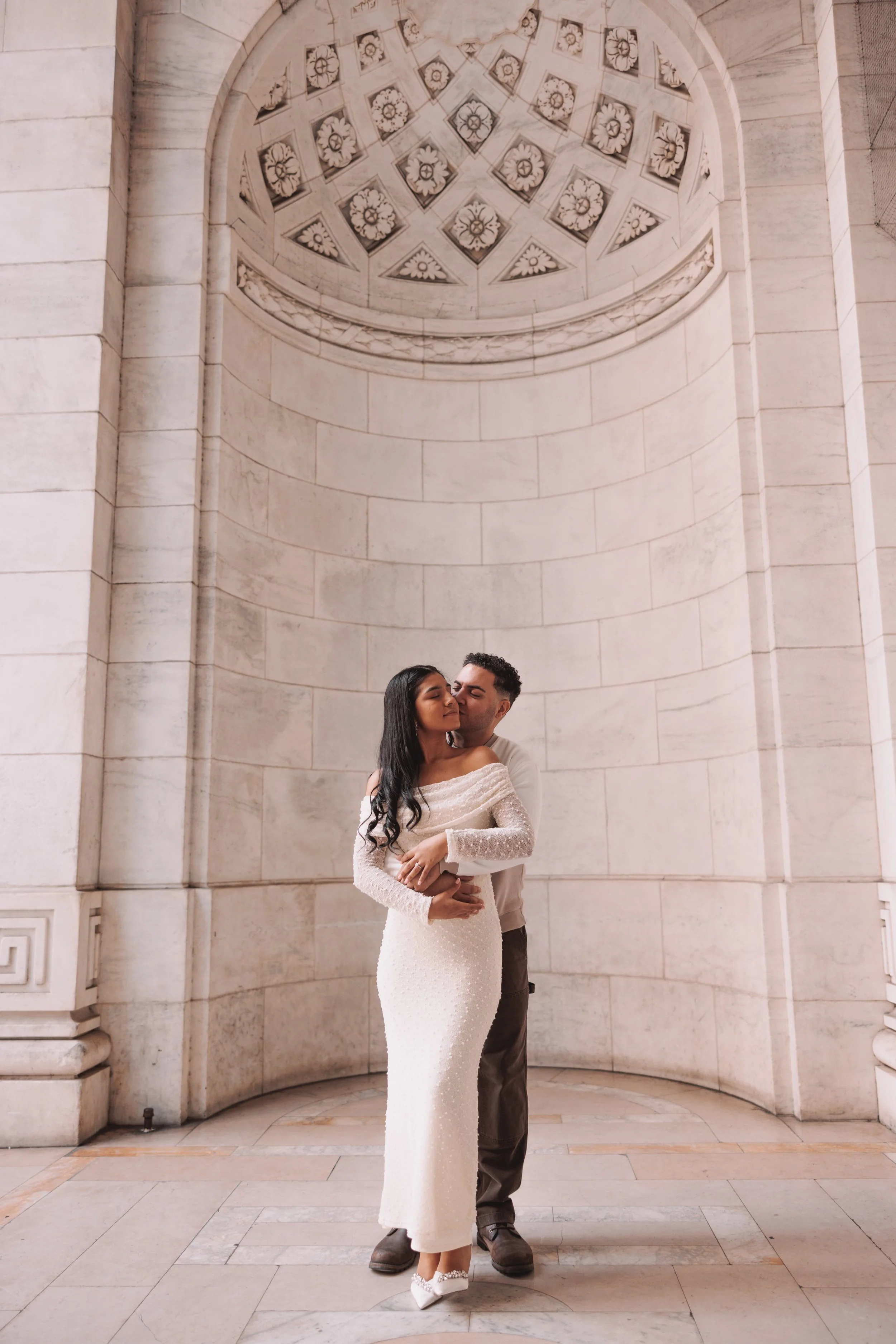 A couple embracing in front of a large, ornate stone wall and a circular ceiling with decorative floral patterns.