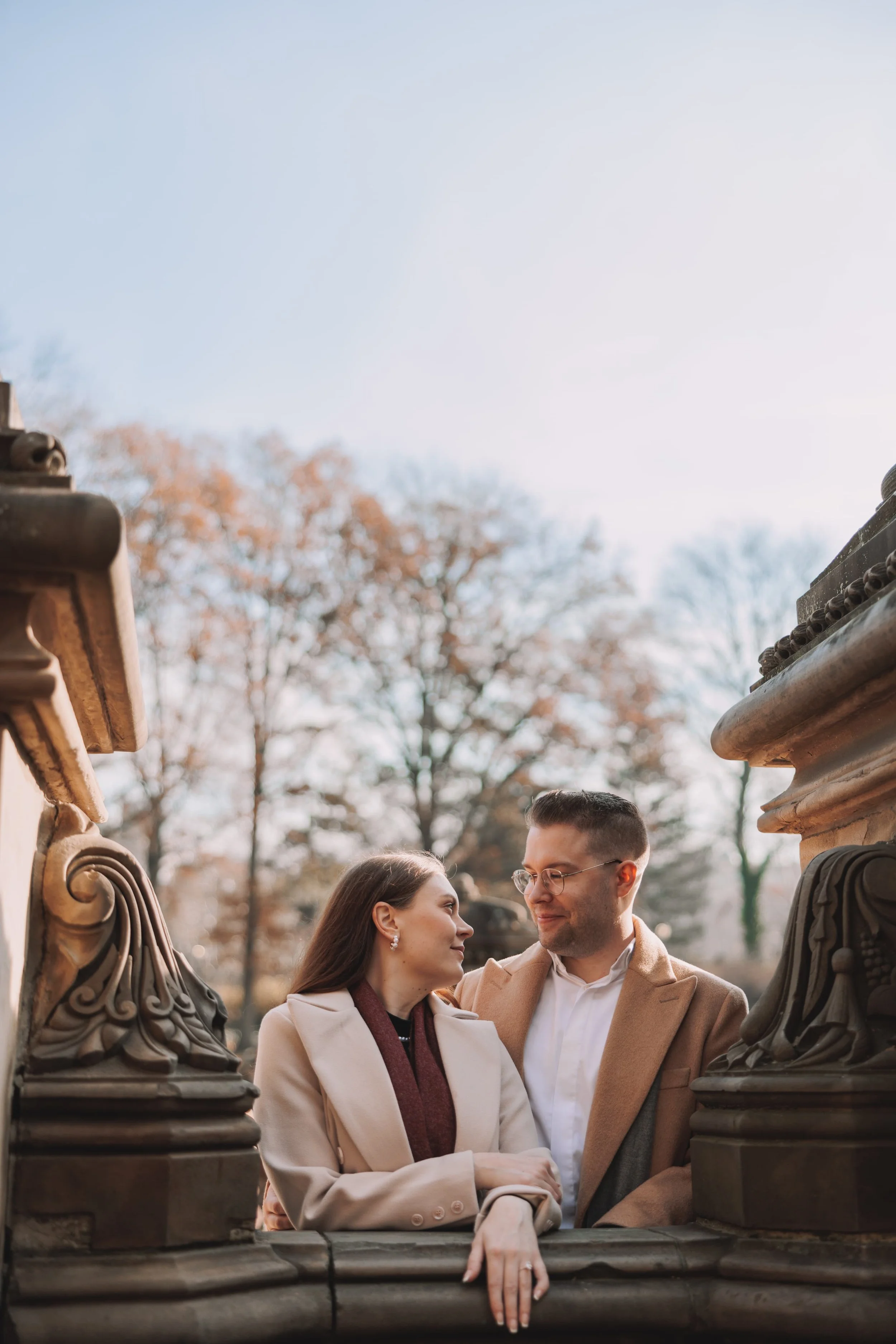 A couple standing outdoors between ornate stone railings, gazing into each other's eyes, with autumn trees in the background.