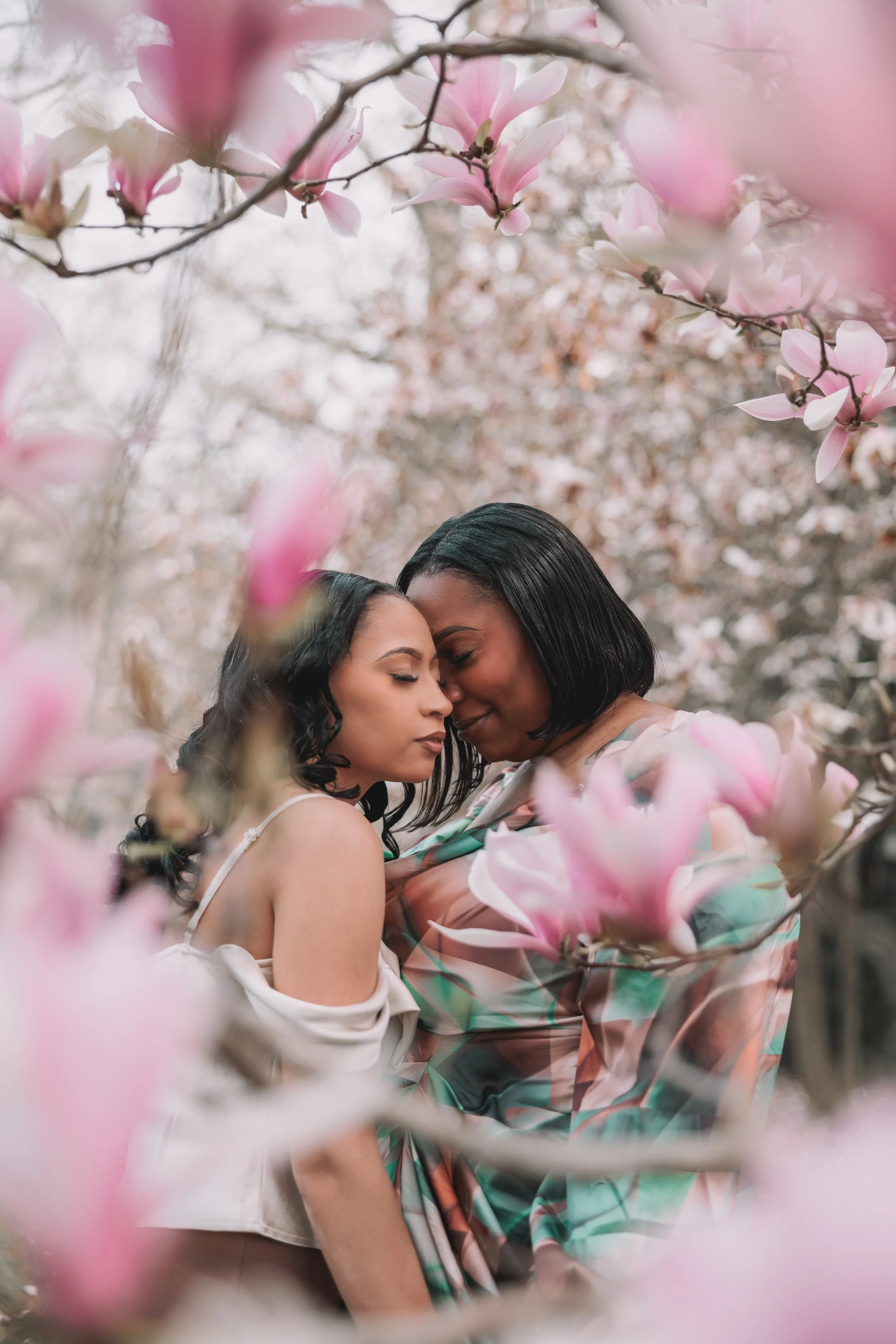 Two women embrace closely amidst blooming pink magnolia flowers on a tree in the background.