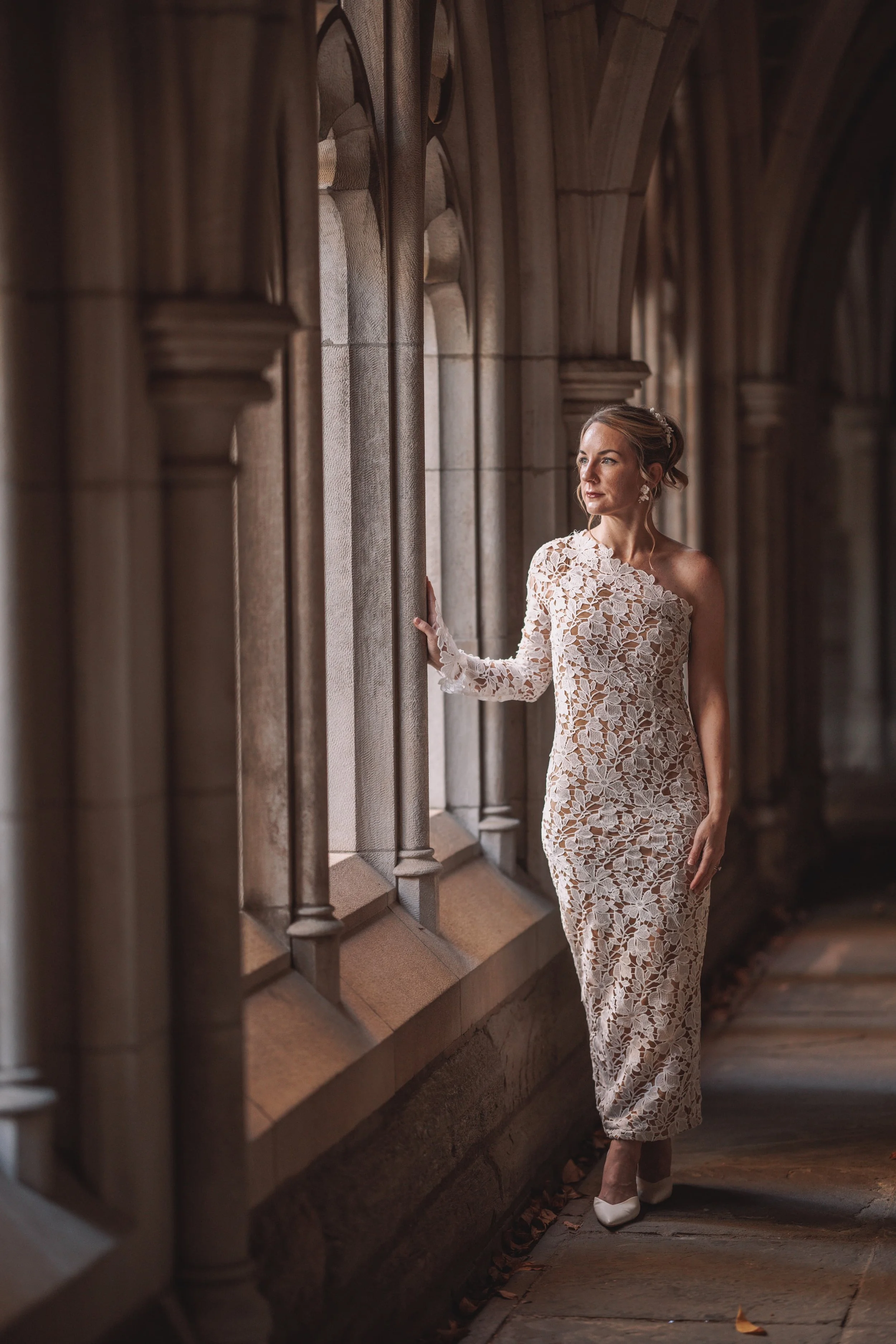 A woman in a white lace dress standing along a stone corridor with arched windows, looking out thoughtfully.