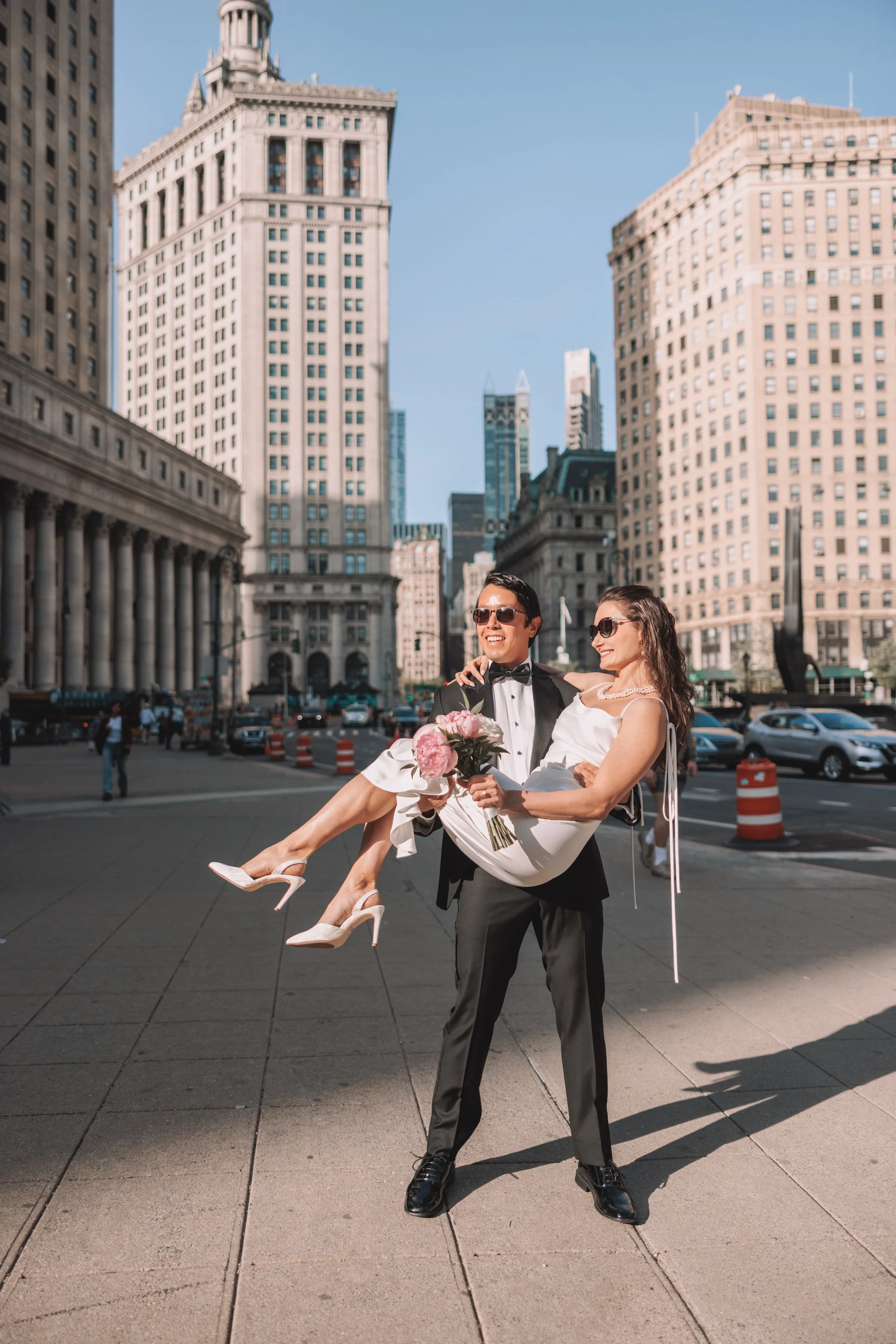 A newlywed couple in wedding attire, the groom in a black tuxedo and the bride in a white dress, smiling and posing in a city street with tall buildings in the background. The groom is holding the bride in his arms, and they appear happy.