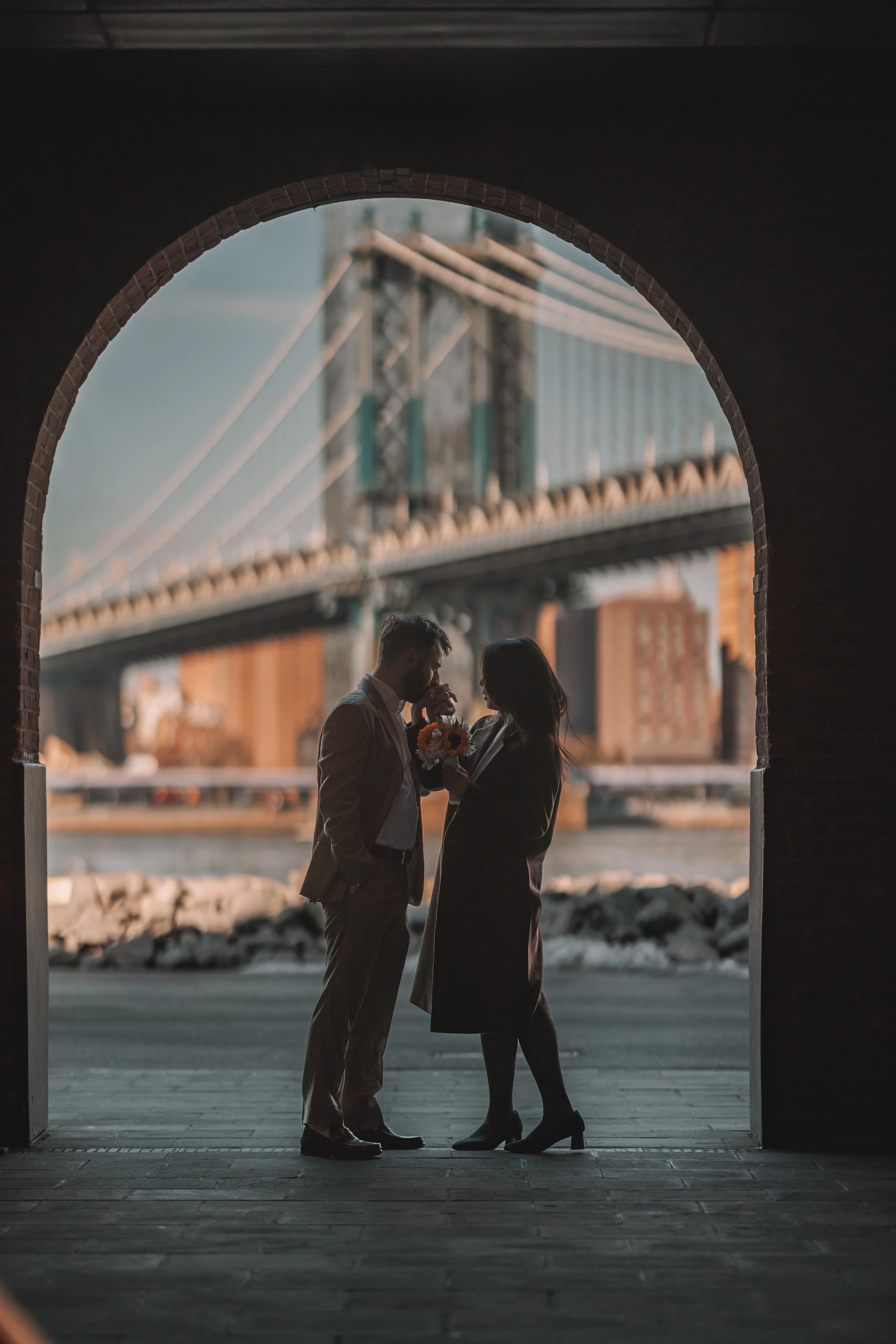 A silhouette of a man presenting flowers to a woman inside a brick archway with a bridge and city skyline in the background.
