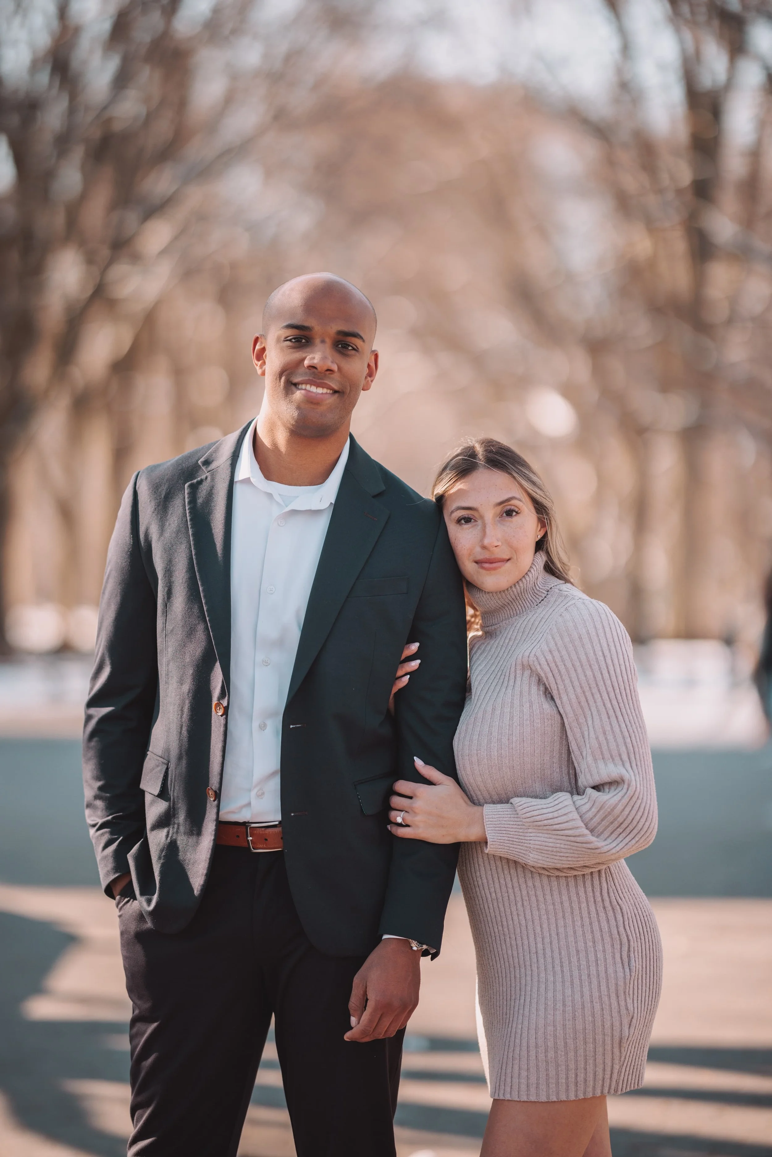 A smiling man in a black suit and white shirt stands outdoors with a woman wearing a beige turtleneck dress, as they pose closely together with her arm around his. The background shows leafless trees and a soft, blurred outdoor setting.