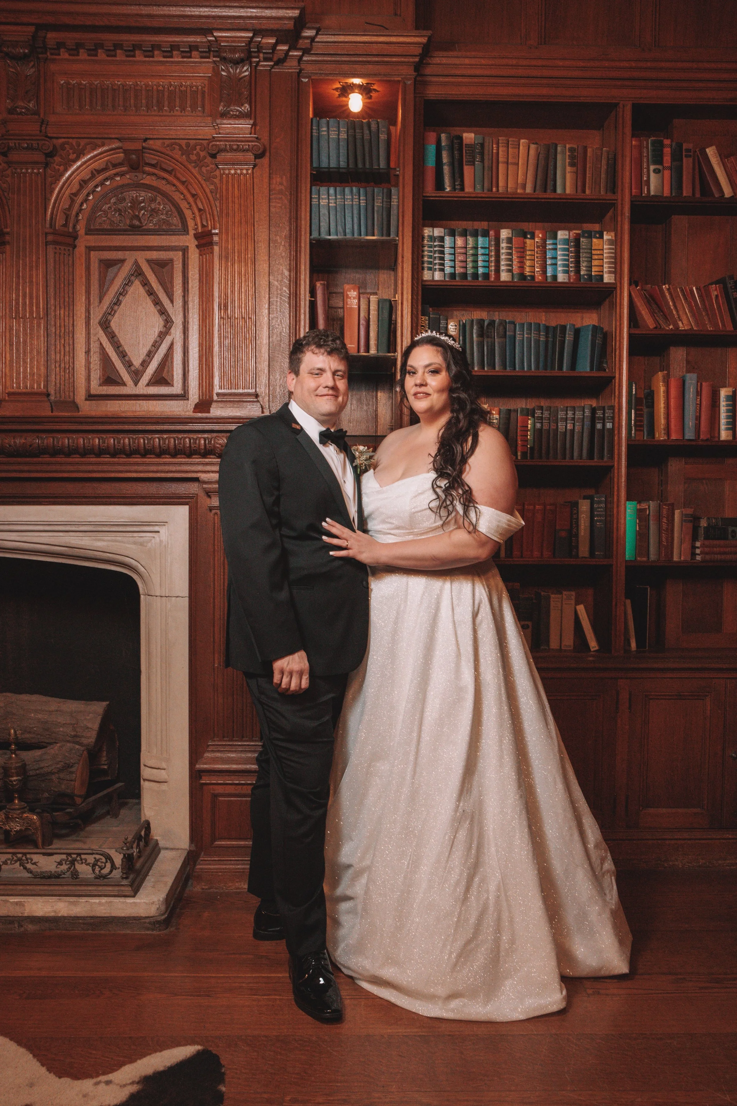 A bride and groom standing together in a wood-paneled room with a fireplace and bookshelves, dressed in wedding attire, smiling.