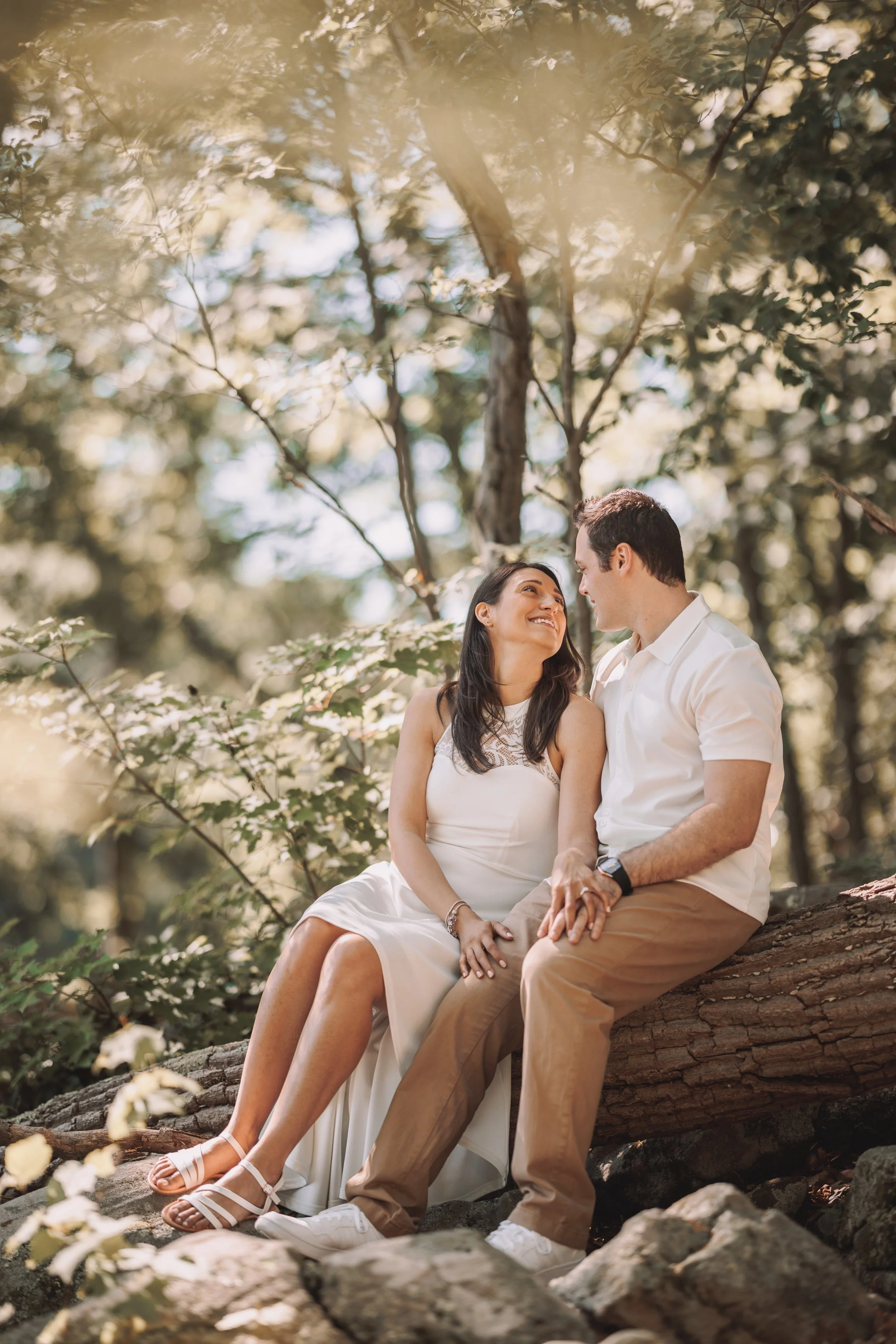 A couple sitting on a fallen tree trunk in a forest, looking at each other and smiling. The woman wears a white dress and the man wears a white shirt and khaki pants.
