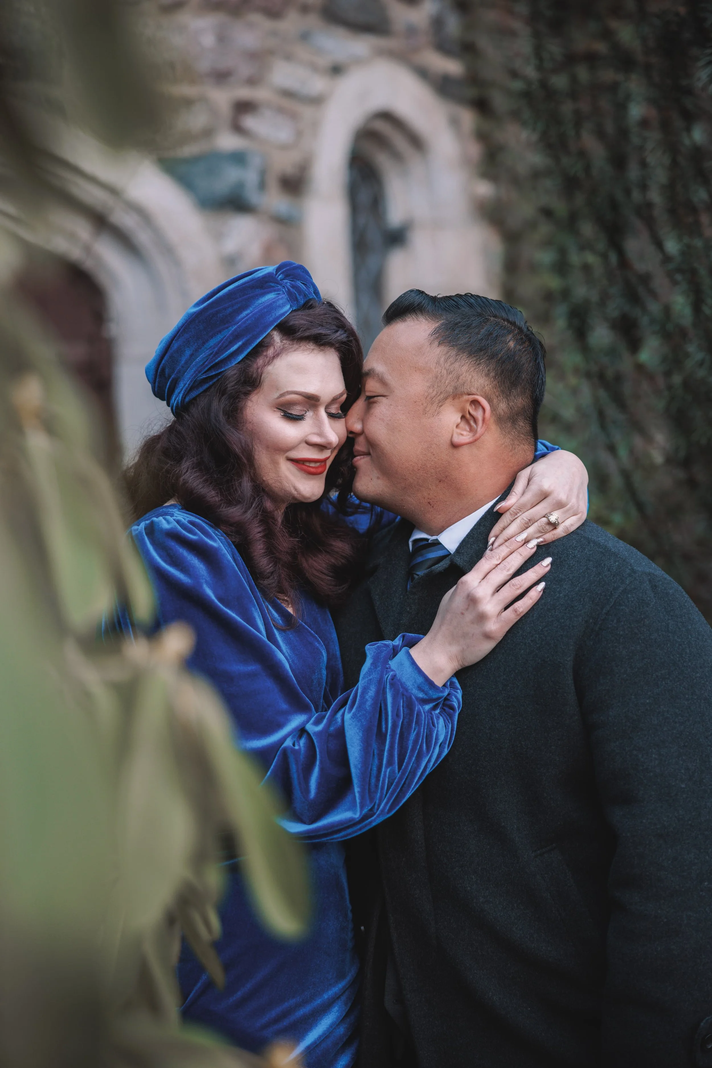 A couple embraces passionately outdoors, with the woman wearing a royal blue velvet dress and matching headscarf, and the man in a dark coat and shirt. They are close together, touching foreheads, with the woman's eyes closed and smiling, and the man