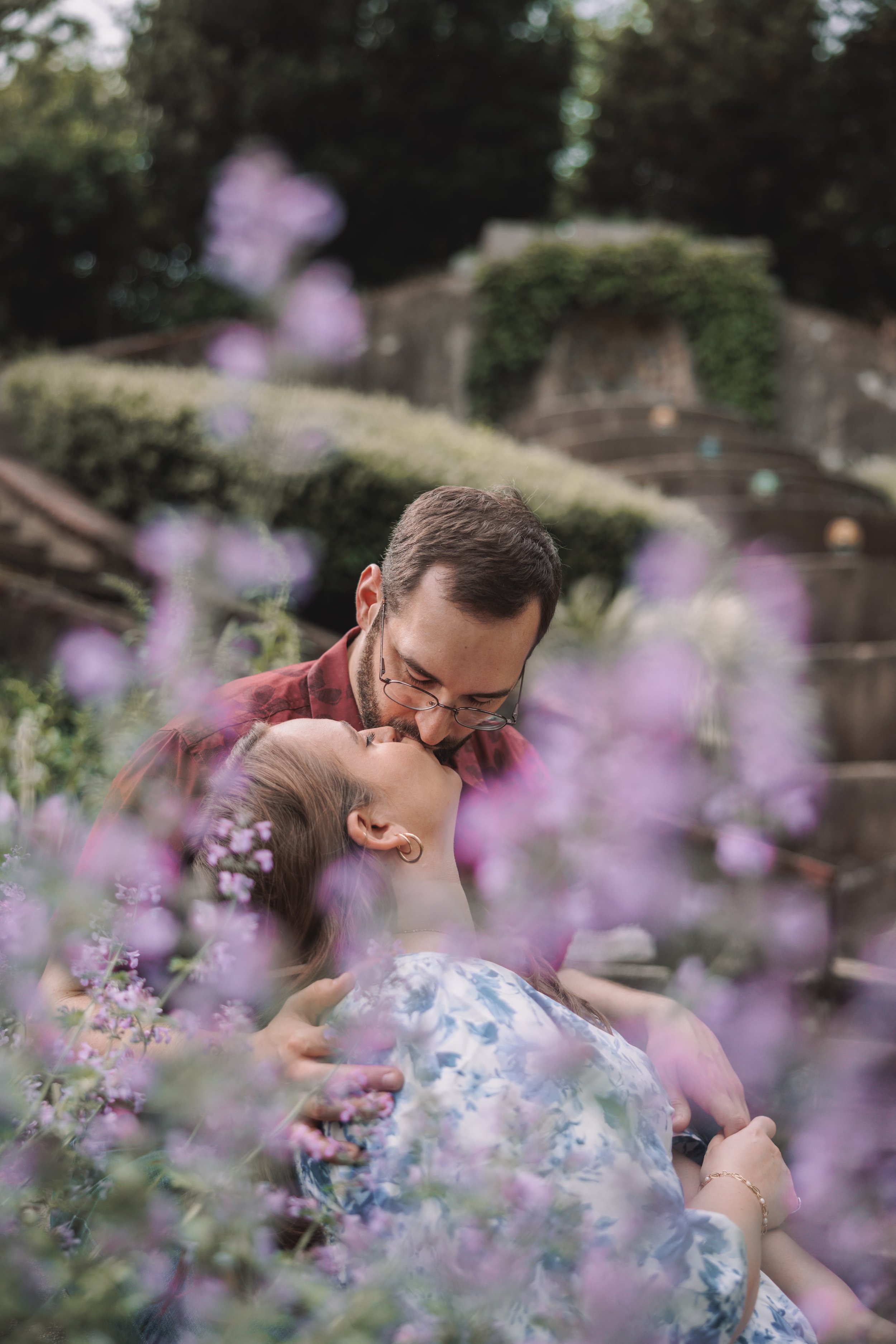 A couple sharing a kiss outdoors, surrounded by purple flowers and greenery.