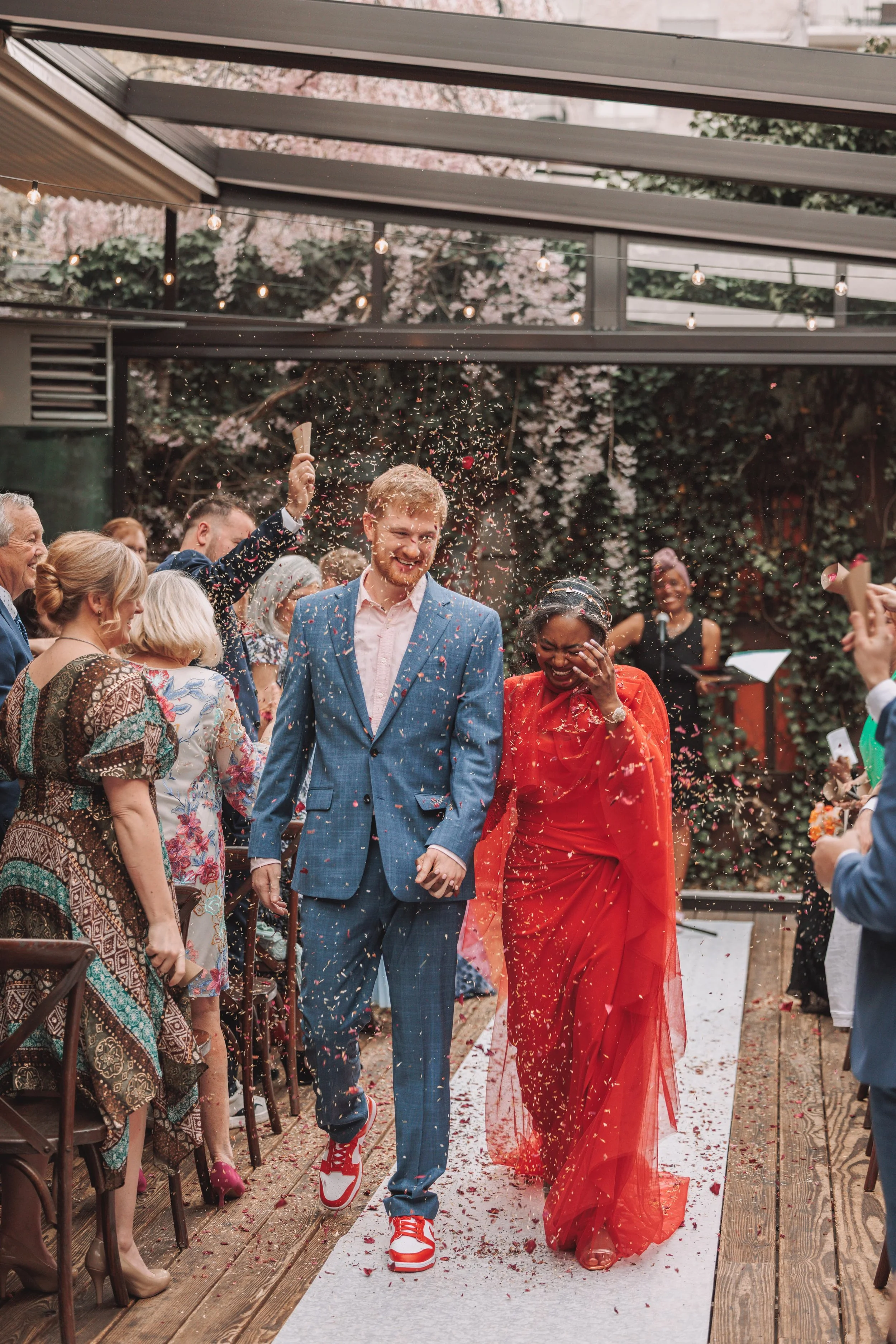 A wedding celebration with a joyful couple walking down the aisle, holding hands, as confetti is thrown around them. The man wears a blue suit with red sneakers, and the woman is in a red dress. Guests are smiling and celebrating under a glass roof w
