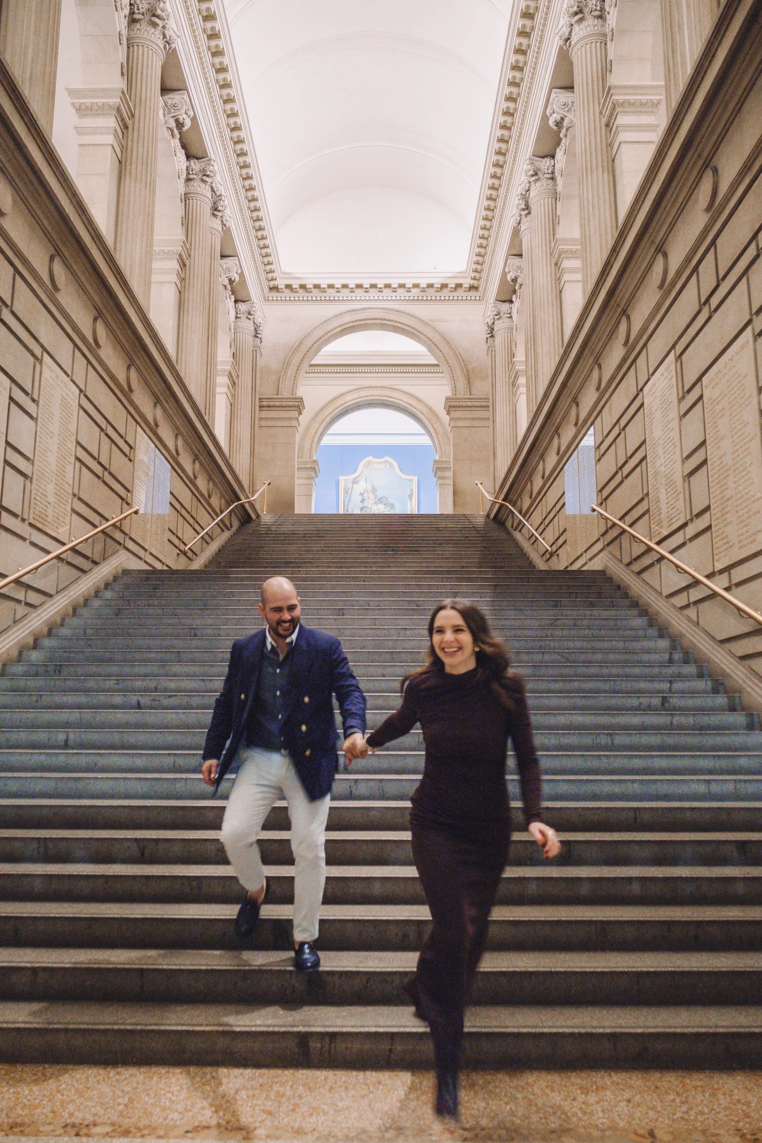 A man and woman happily running down a staircase inside a grand, historic building with ornate architectural details.