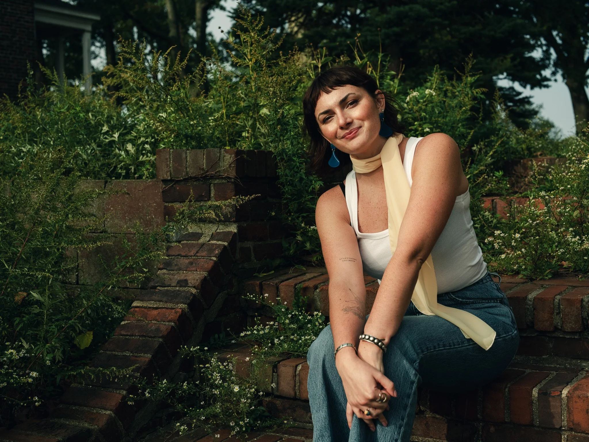 A woman with dark hair and bangs sits on a brick ledge in a garden, smiling at the camera. She is wearing a white tank top, blue jeans, and blue earrings, with a light yellow scarf around her neck.