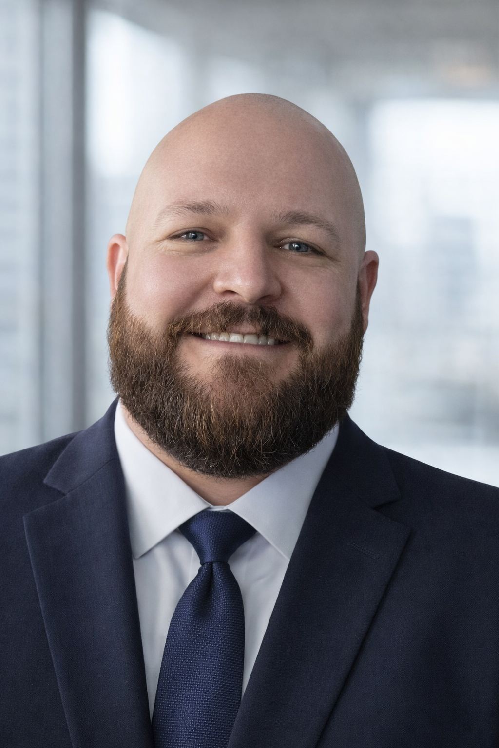 A smiling man with a beard and bald head, wearing a dark suit, white shirt, and dark tie, in a professional setting.