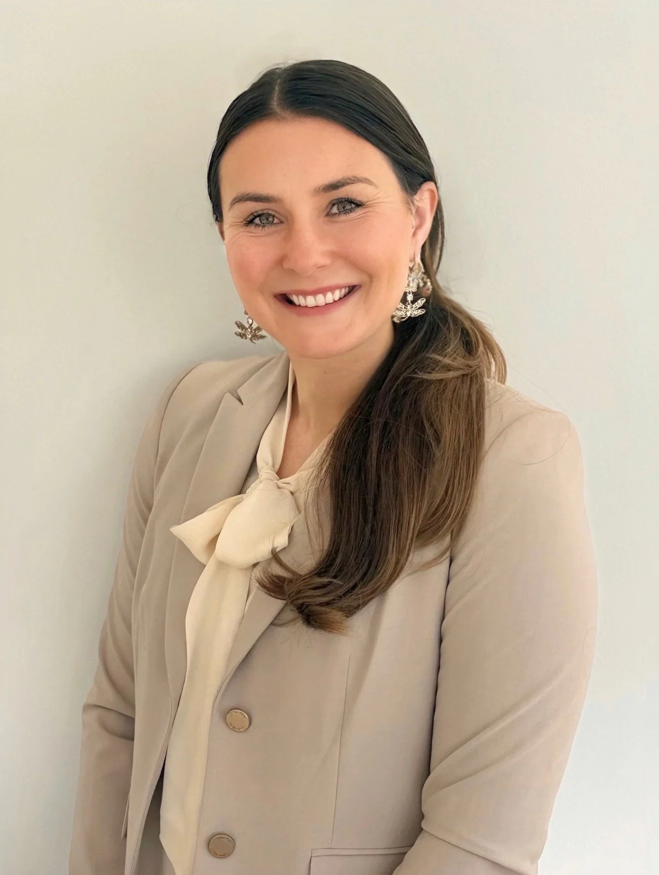 Professional executive headshot of Kelly McCabe smiling, wearing a beige blazer and cream blouse with decorative earrings.