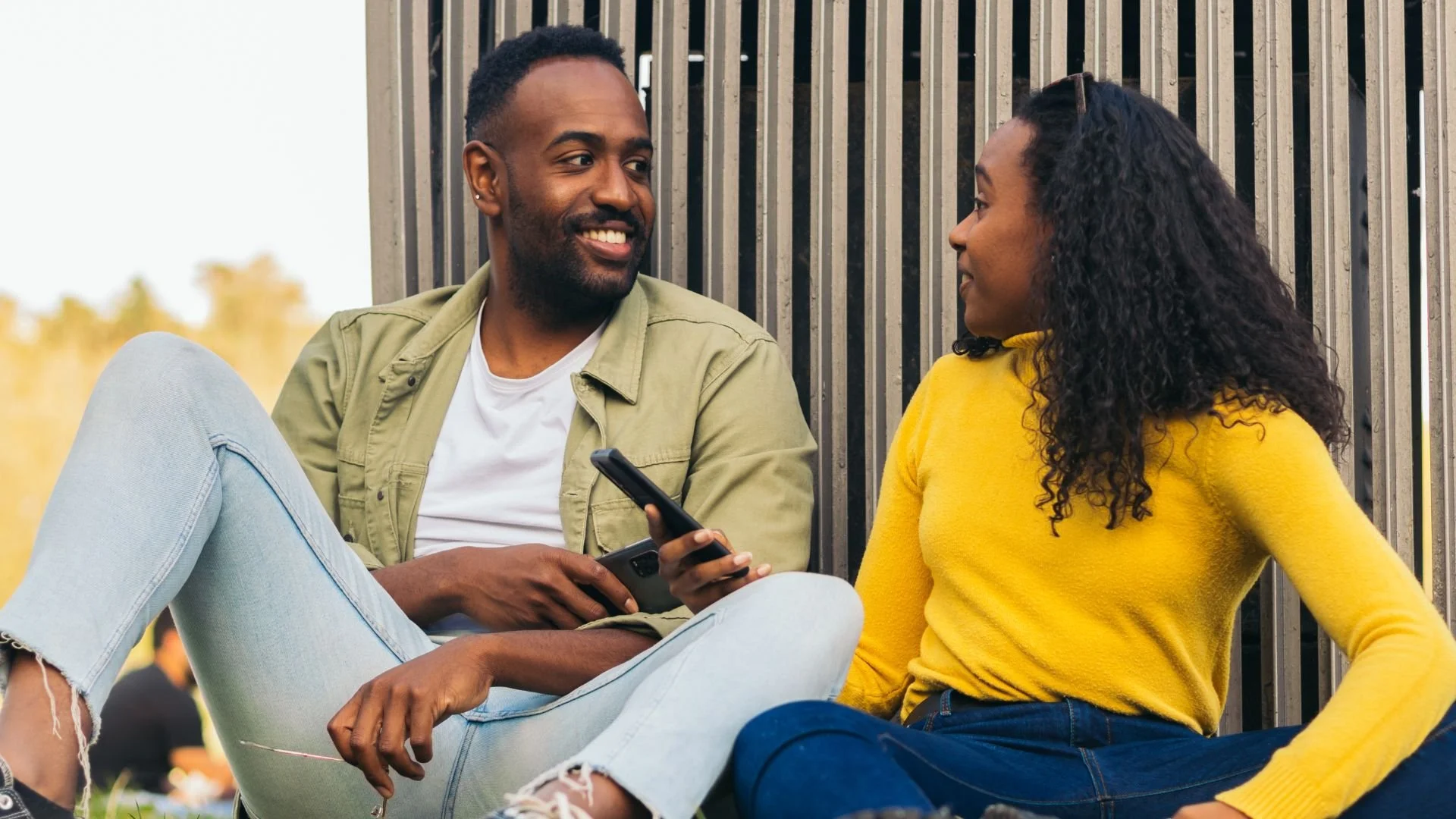 A Black couple engaged in conversation while sitting outdoors during the day in a bright, natural environment.