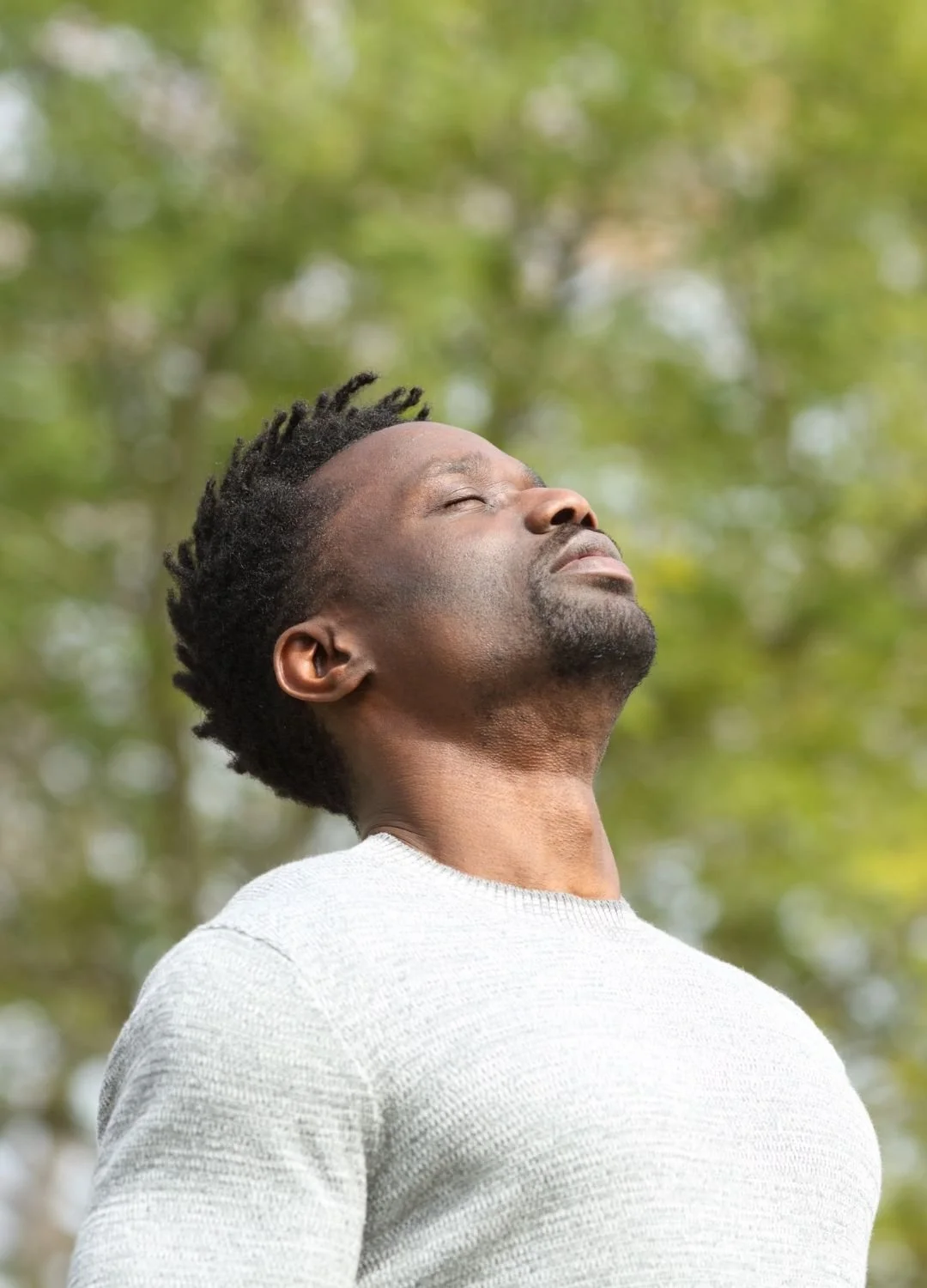 A serious Black man standing in a lush green park, eyes closed, taking a deep breath of fresh air to practice mindfulness.