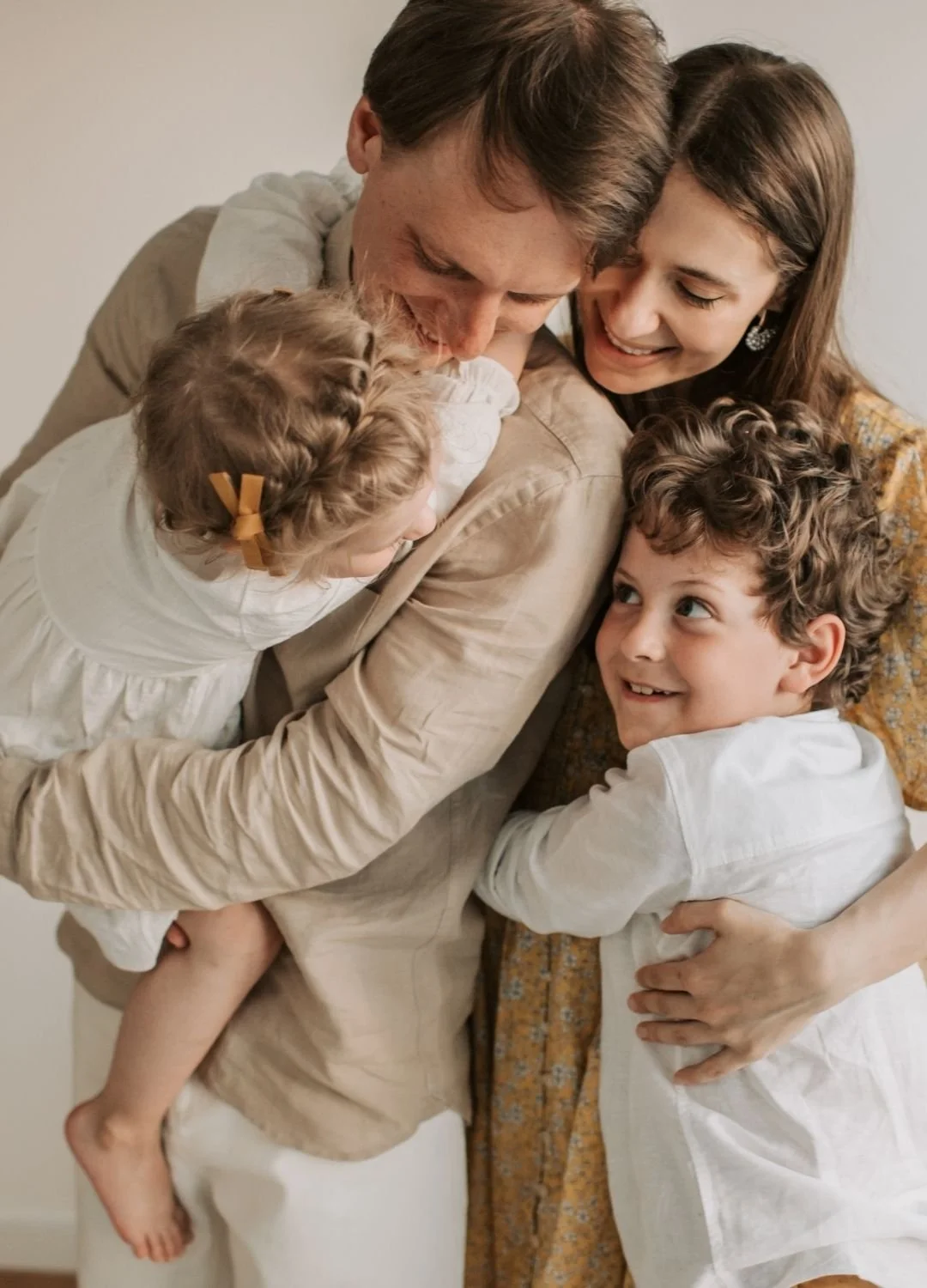 A candid indoor portrait of a family of four sharing a group hug, showing a father holding a toddler and a mother embracing her young son.