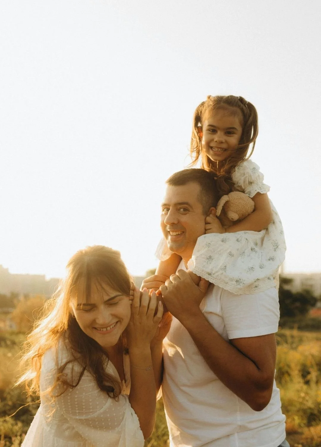 A happy family of three during a golden hour outdoor session, featuring a father carrying his young daughter on his shoulders while the mother smiles beside them.