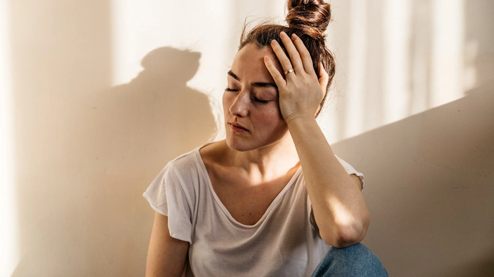 Close-up of a woman with her eyes closed, holding her hand to her forehead while suffering from a headache or migraine in a sunlit room.