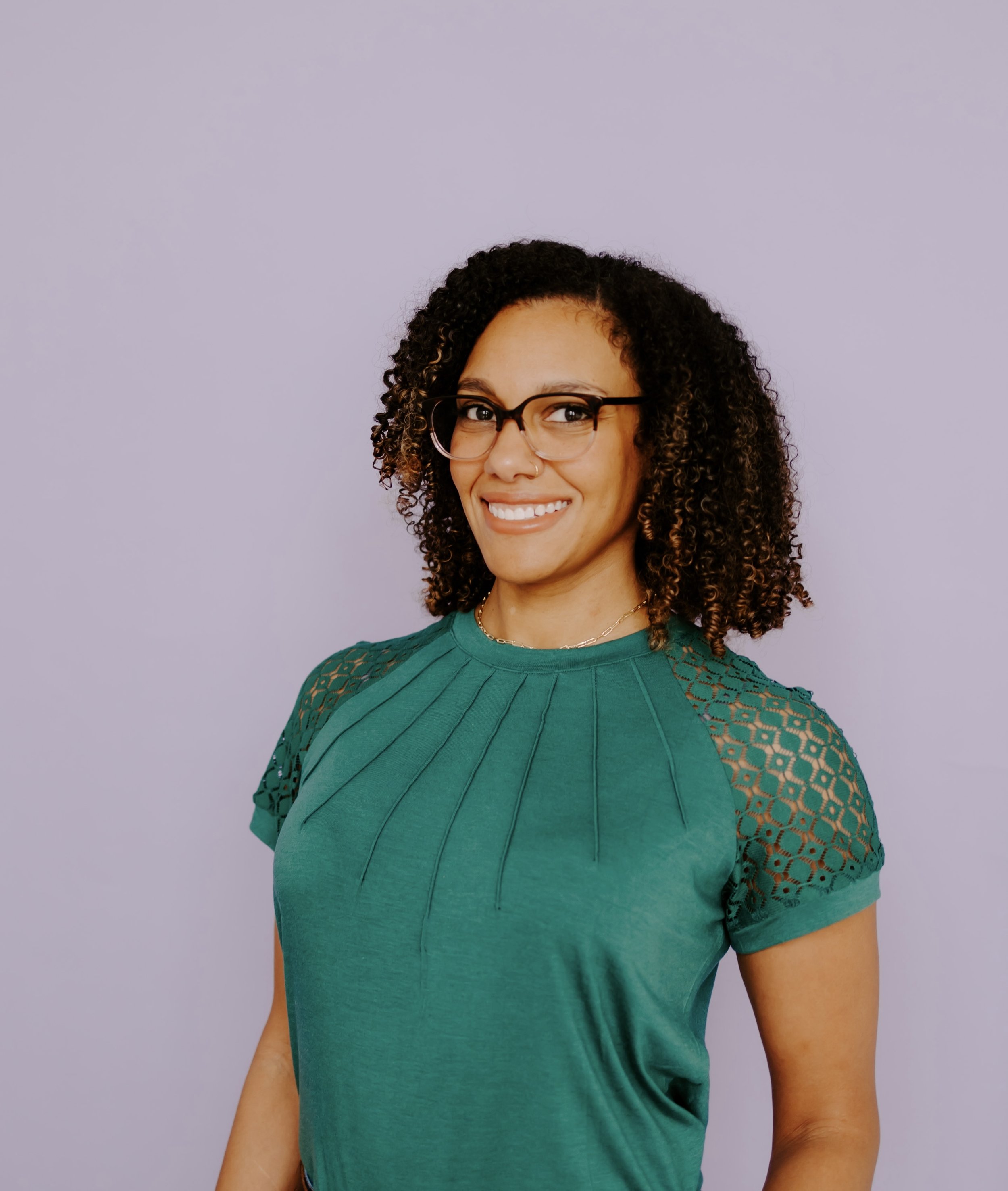 A smiling woman with curly hair, glasses, and a nose piercing, wearing a teal top with lace sleeves, standing against a plain light purple background.