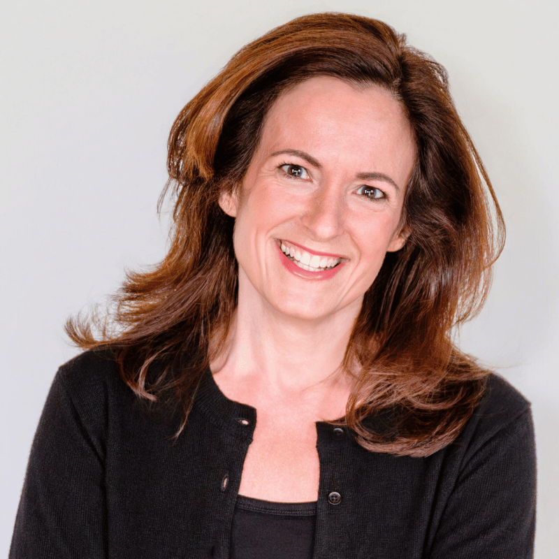 A woman with reddish-brown hair smiling, wearing a black top and black cardigan, against a plain light background.